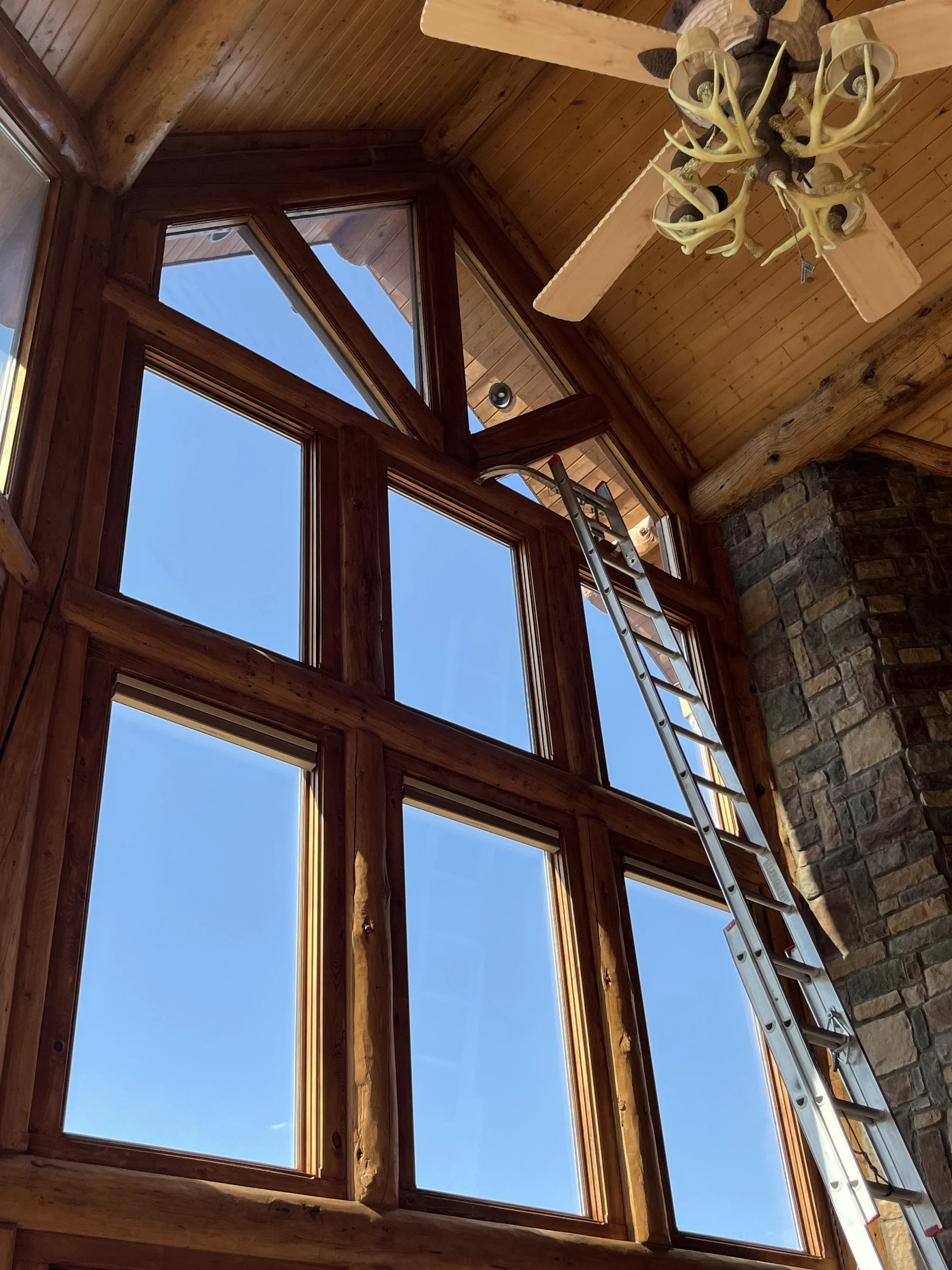 A room with large wooden framed windows showing a clear blue sky outside; a ladder leaning against the window wall, and a ceiling mounted antler chandelier.