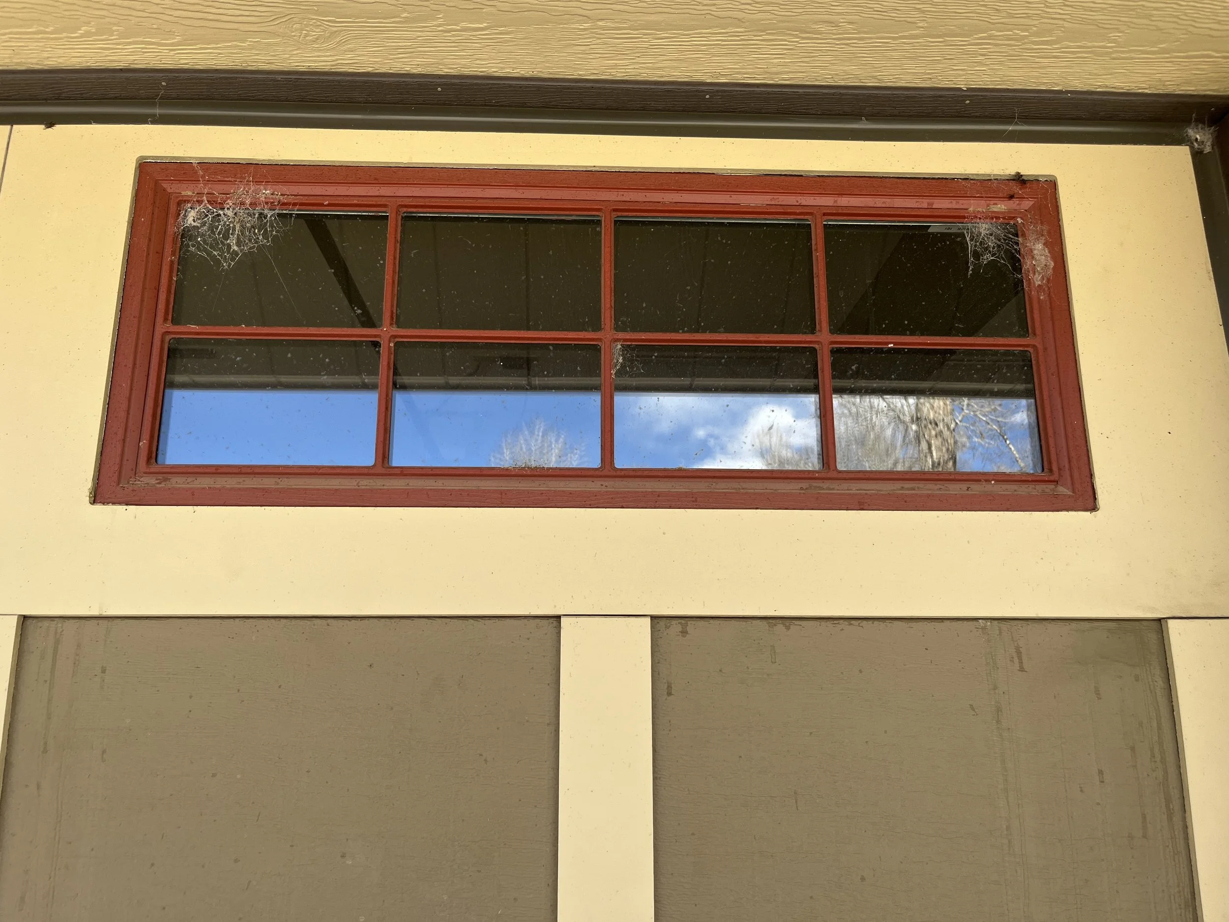 A large, rectangular window with a red frame, divided into smaller panes, set into a yellow wall with black trim at the top. The window shows a reflection of a blue sky with white clouds and leafless trees. There are cobwebs in the corners of the win