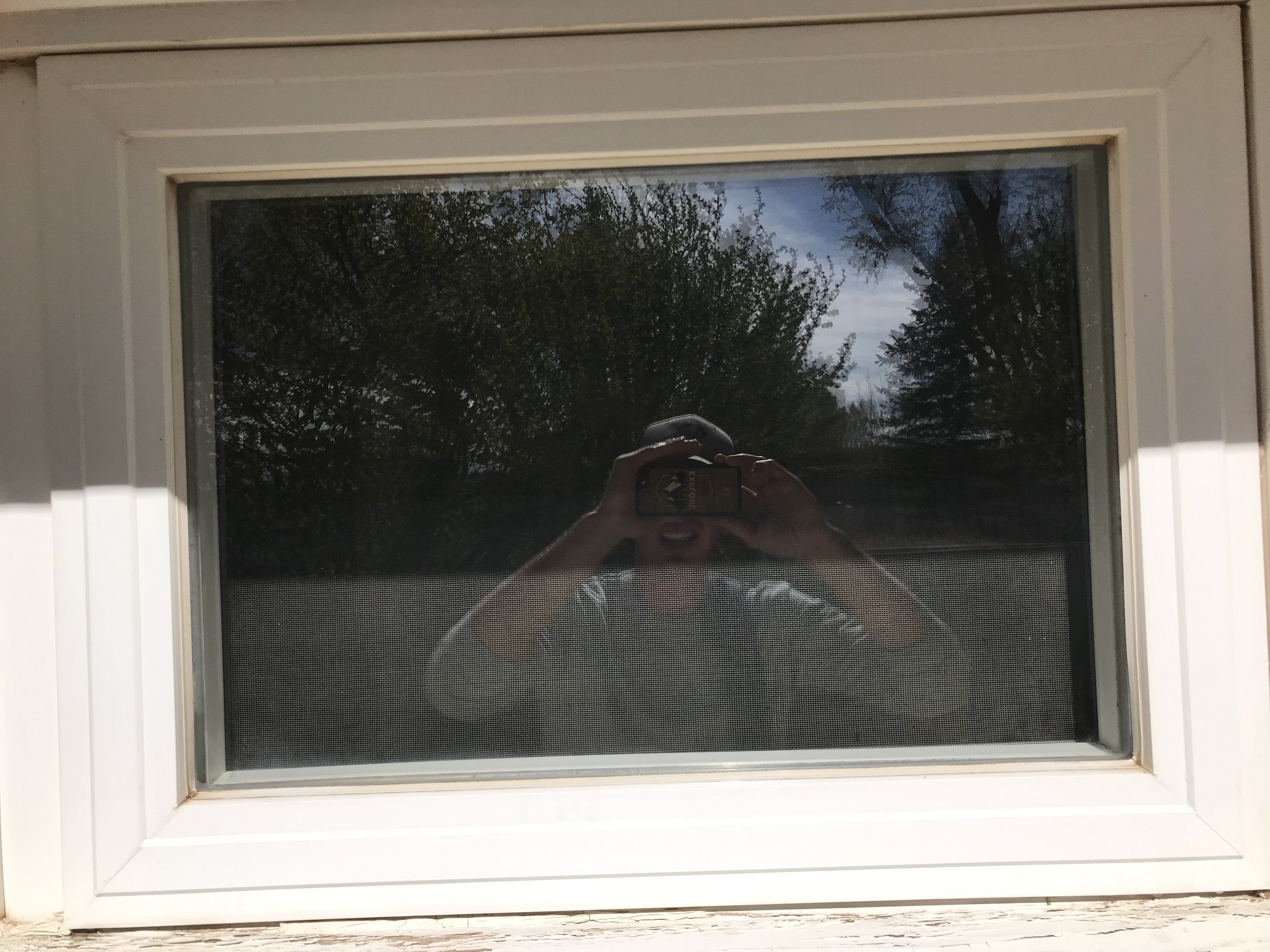 Outside view of a house window with a reflection of the trees and sky in the glass, and a person taking the photo visible in the reflection.