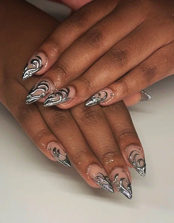 Person with long, metallic silver and black artistic nail designs on fingernails, hands resting on a white surface.