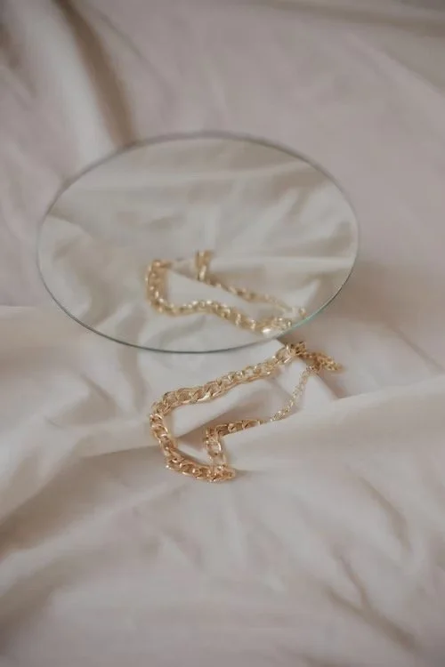 A gold chain necklace resting on a white fabric with a small round mirror reflecting part of the chain.