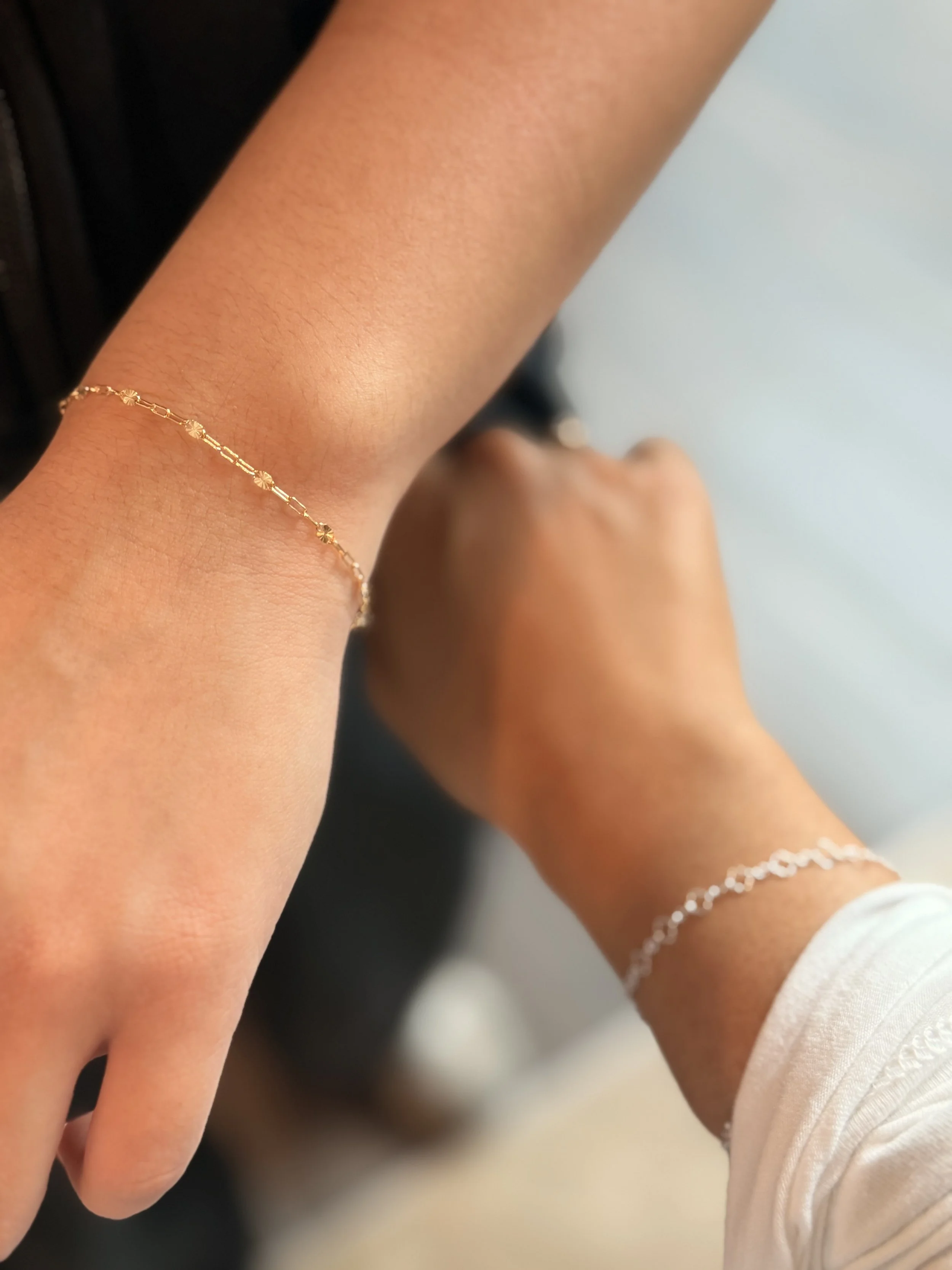 Close-up of a woman's hand and wrist with a delicate gold bracelet with small round and rectangular links, wearing a white long-sleeved top.