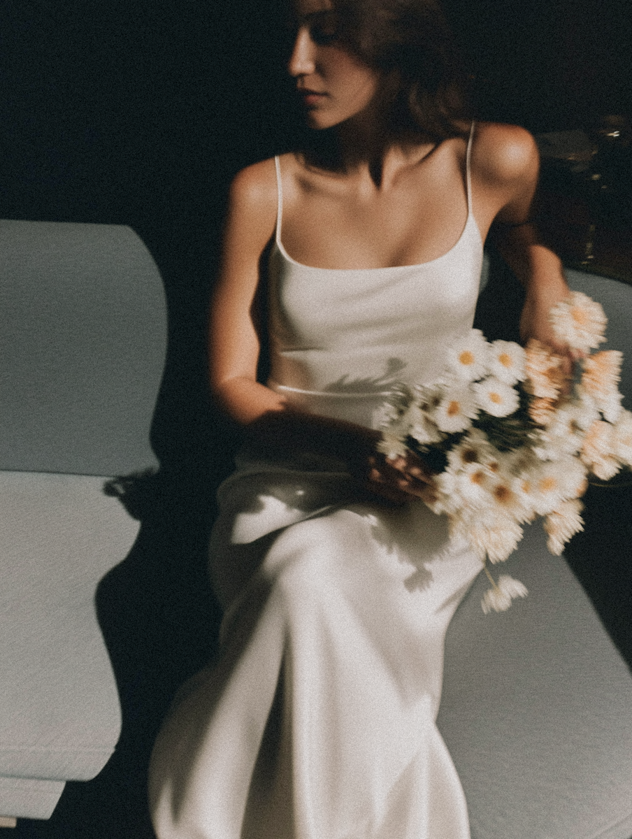 A woman sits on a gray bench holding a bouquet of white daisies, dressed in a white satin slip dress.