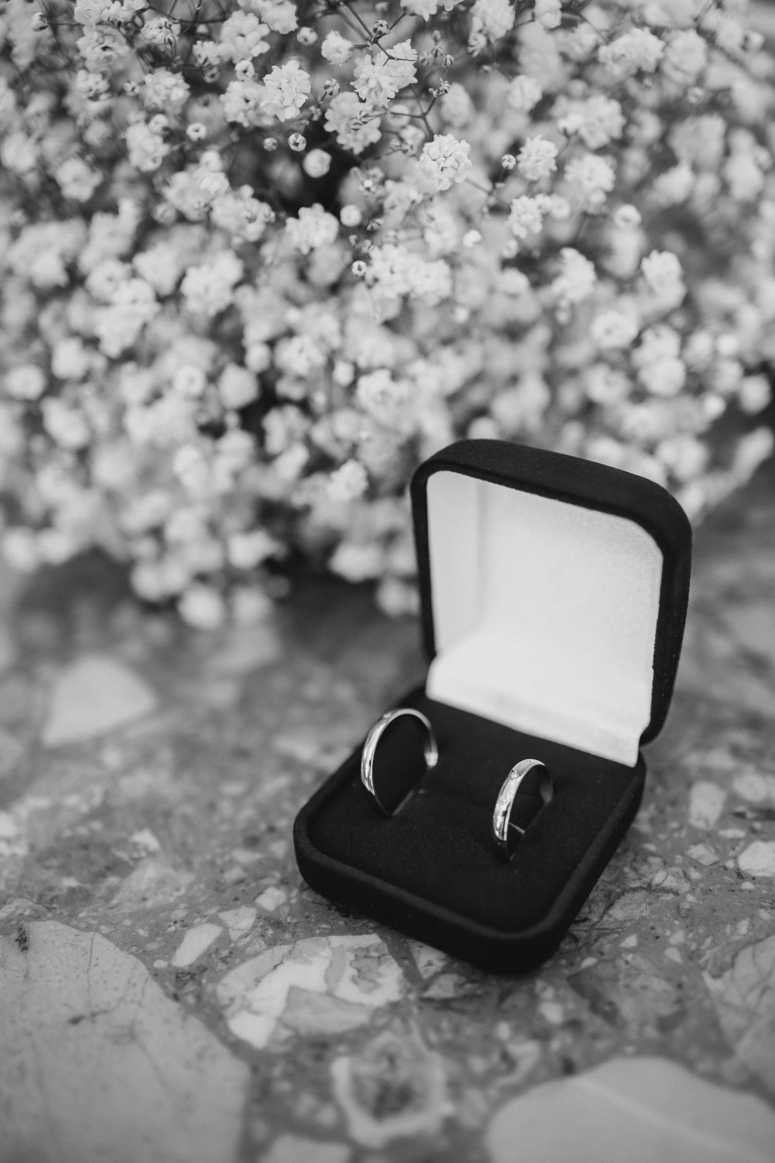 A black jewelry box with a pair of wedding rings inside, placed on a granite surface; in the background, there is a bouquet of small white flowers.