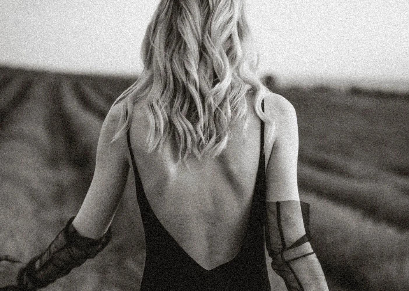 Black and white photograph of a woman with wavy blonde hair, wearing black lingerie, with her back to the camera, and her face not visible.