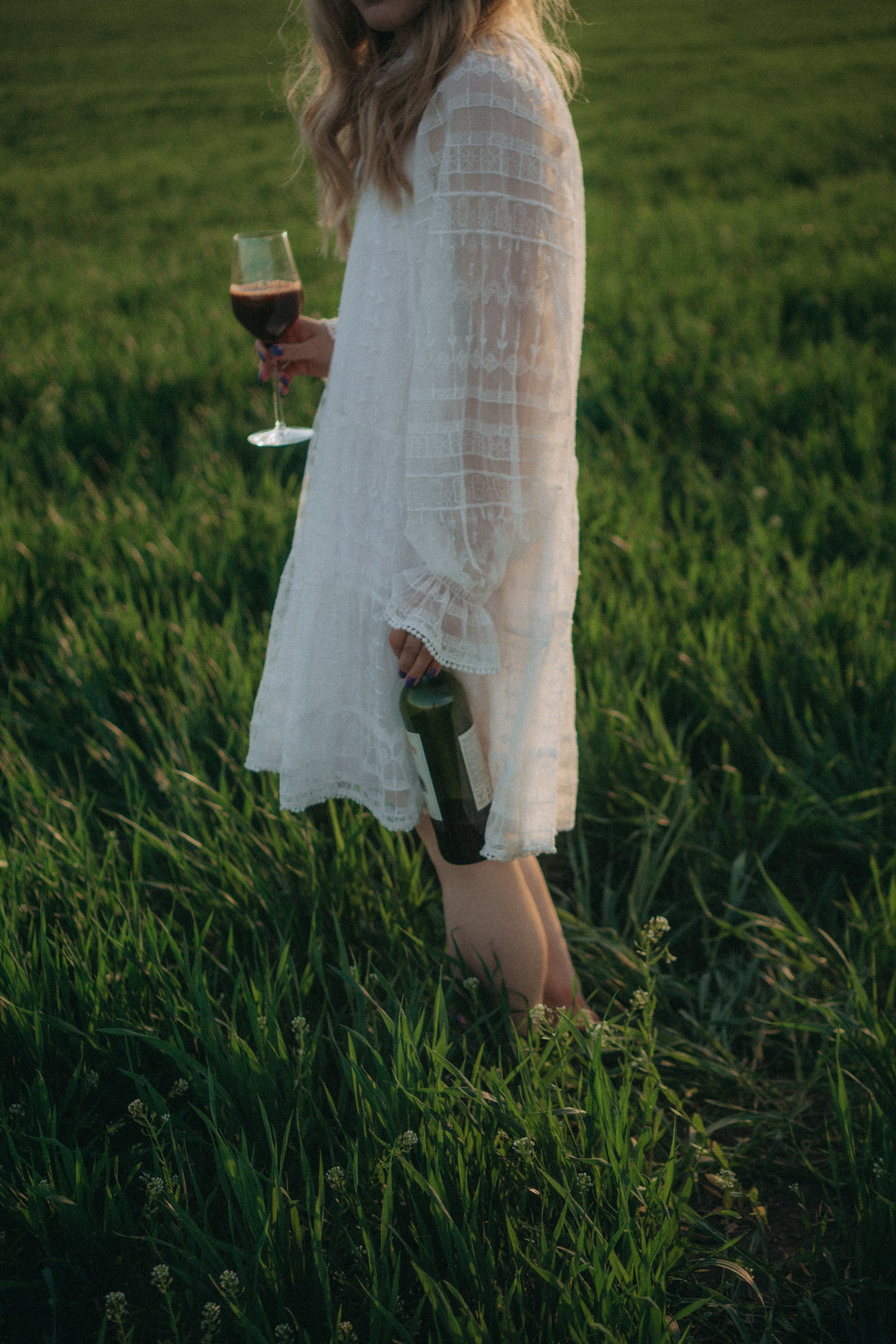 A woman wearing a white lace dress standing in a grassy field, holding a glass of red wine in her right hand and a wine bottle in her left.