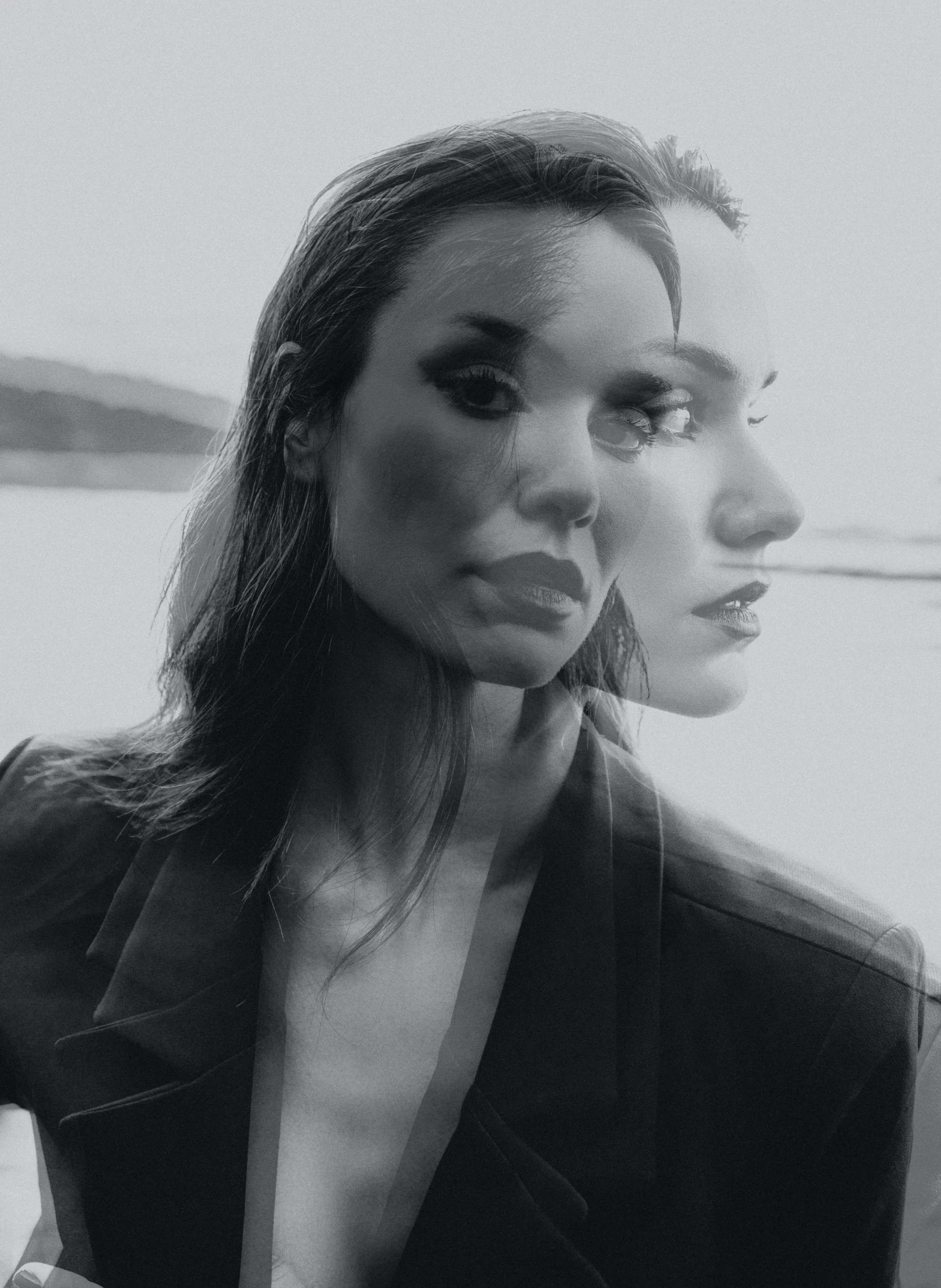 A black and white photo of a woman with voluminous hair, wearing dark makeup, and a blazer over a white top, with a blurred natural landscape in the background.