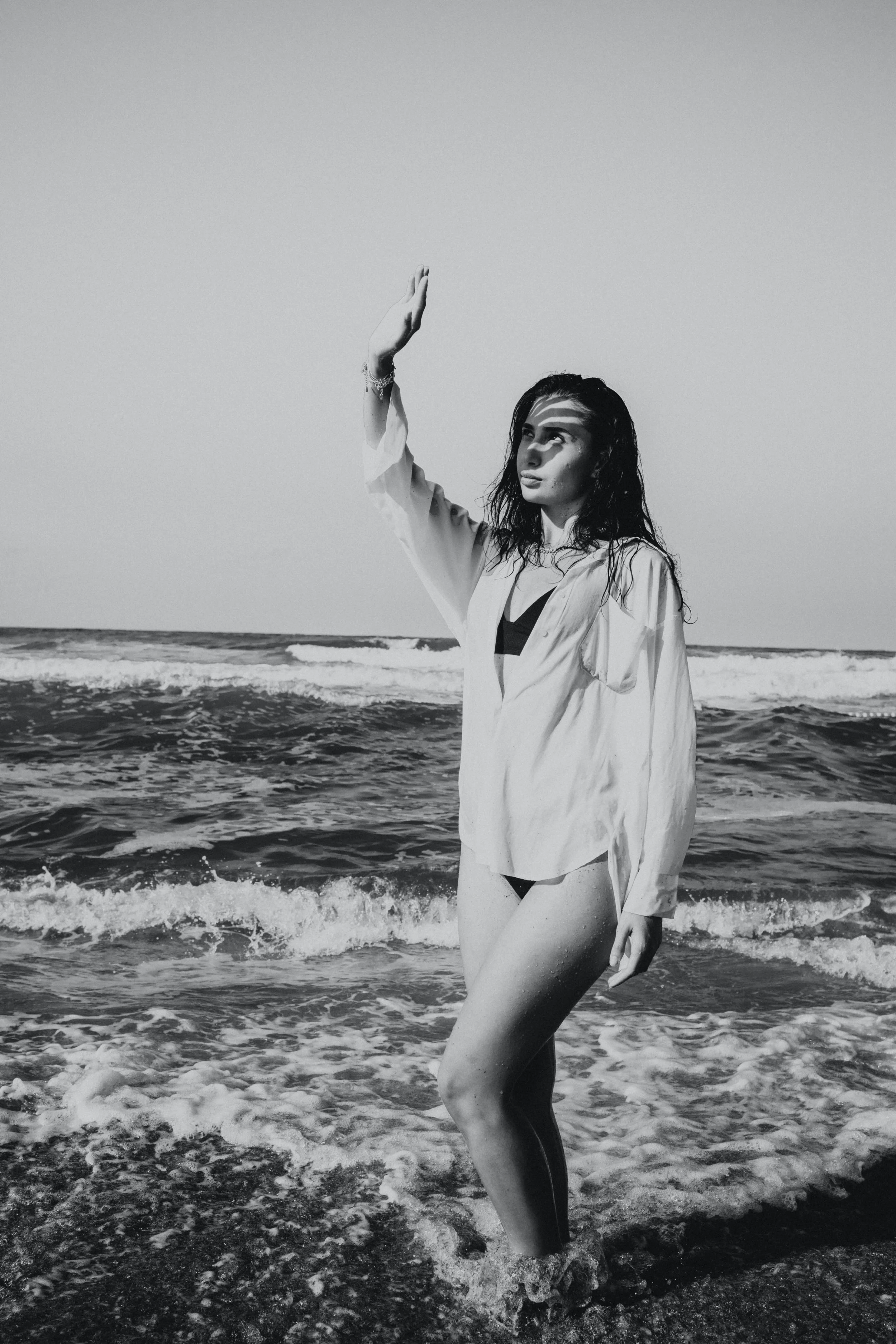 Black and white photo of a woman standing in the ocean waves, raising one hand, with wet hair and light clothing.