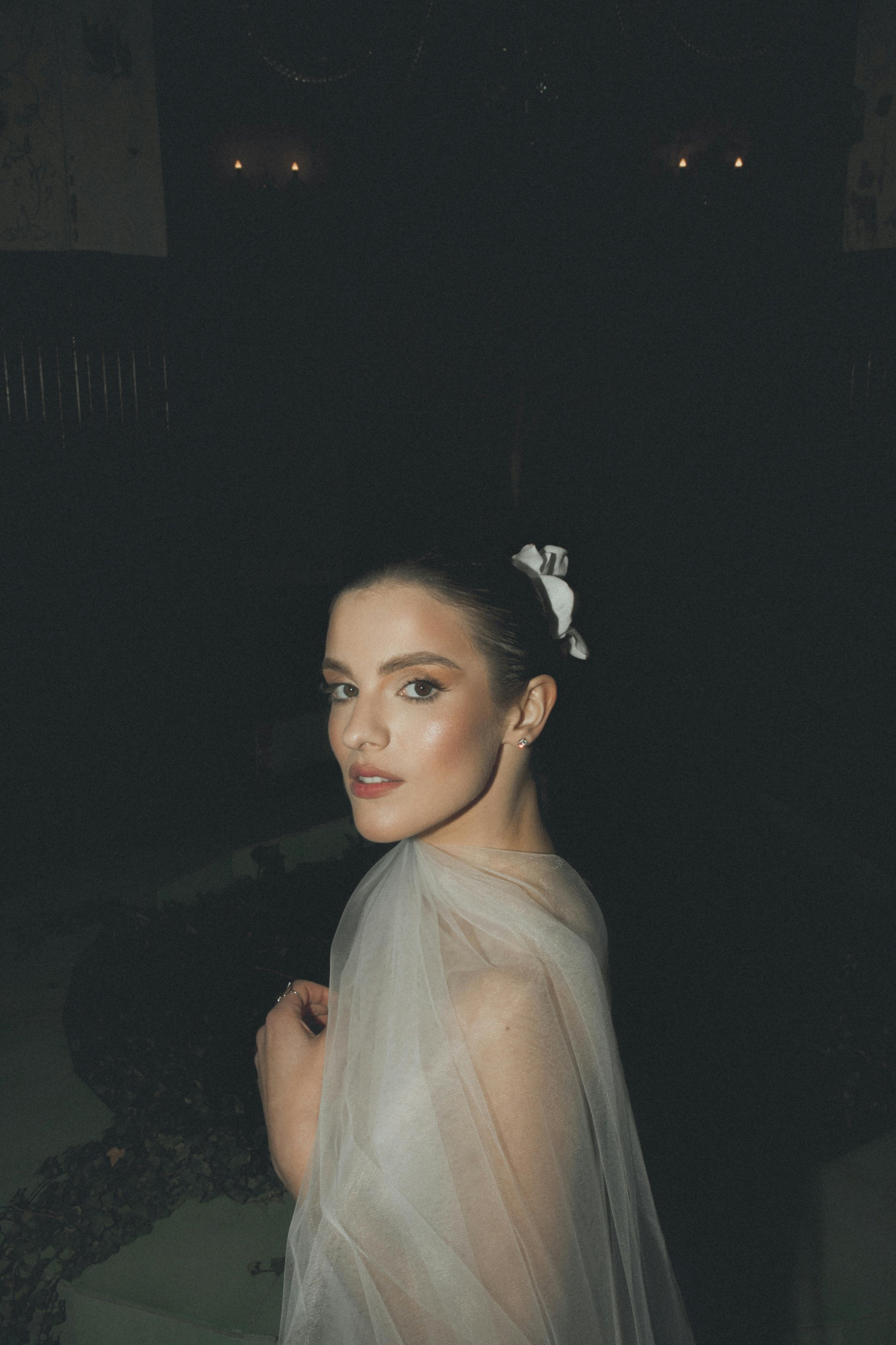 A young woman with dark hair pulled back, wearing a white or light-colored sheer dress, with a white bow in her hair, looking over her shoulder in a dimly lit setting.