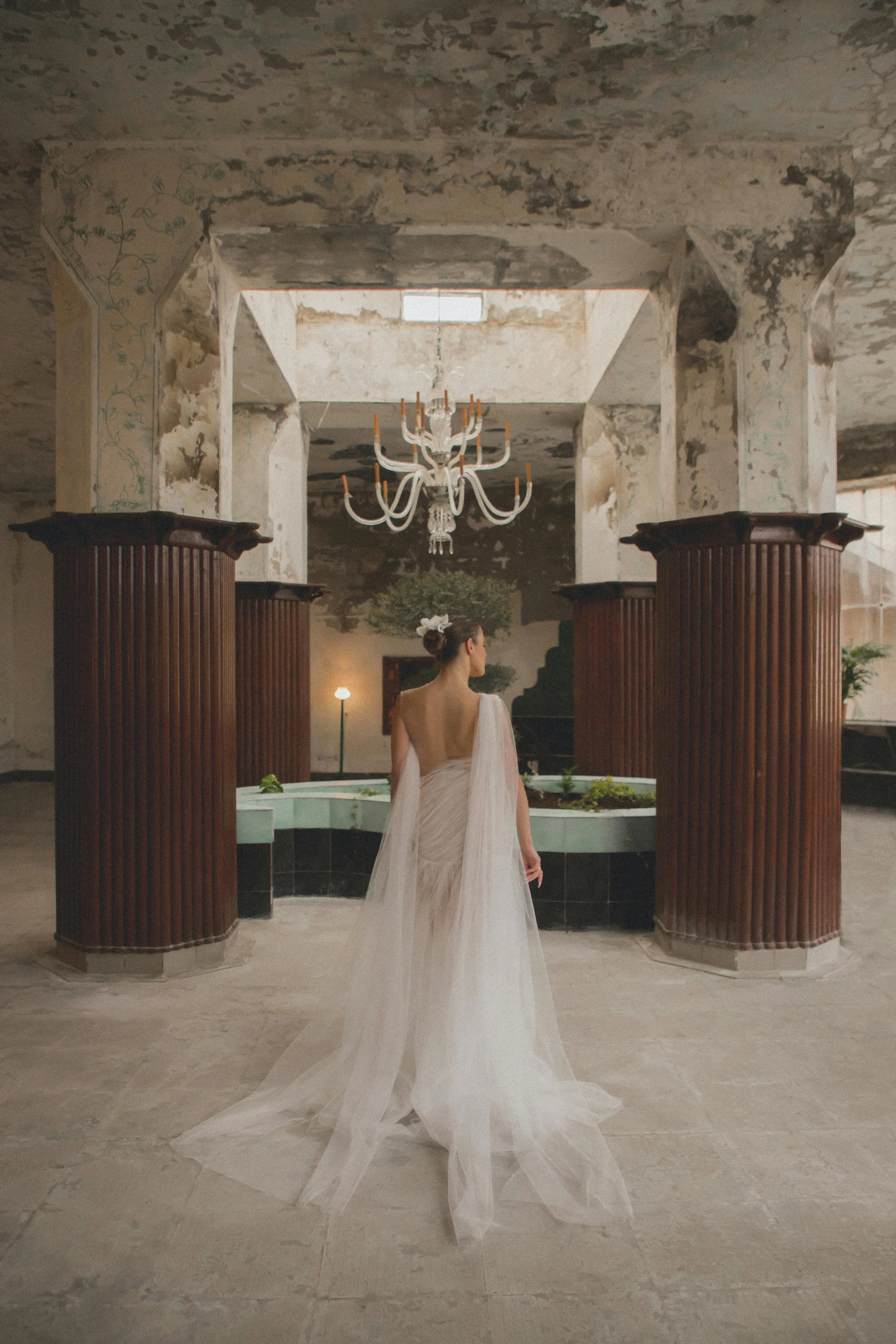 A woman in a white wedding dress standing inside a historic, rustic building with peeling paint, a chandelier, and wooden columns.