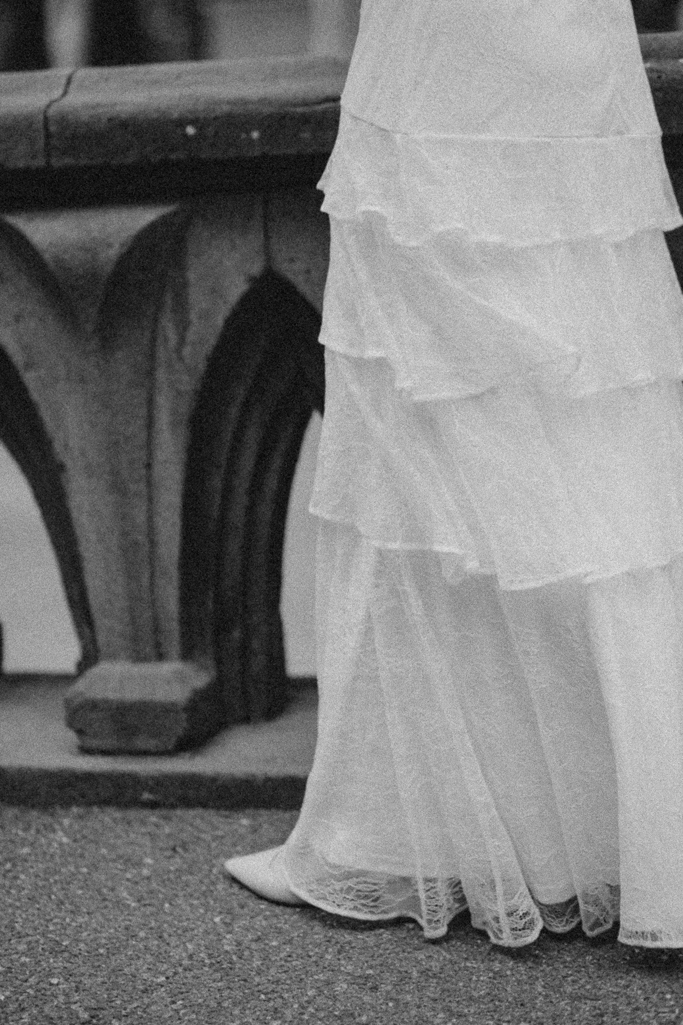 Close-up of the lower part of a person wearing a wedding dress with lace details and high heels, standing on a textured floor near a wood table.