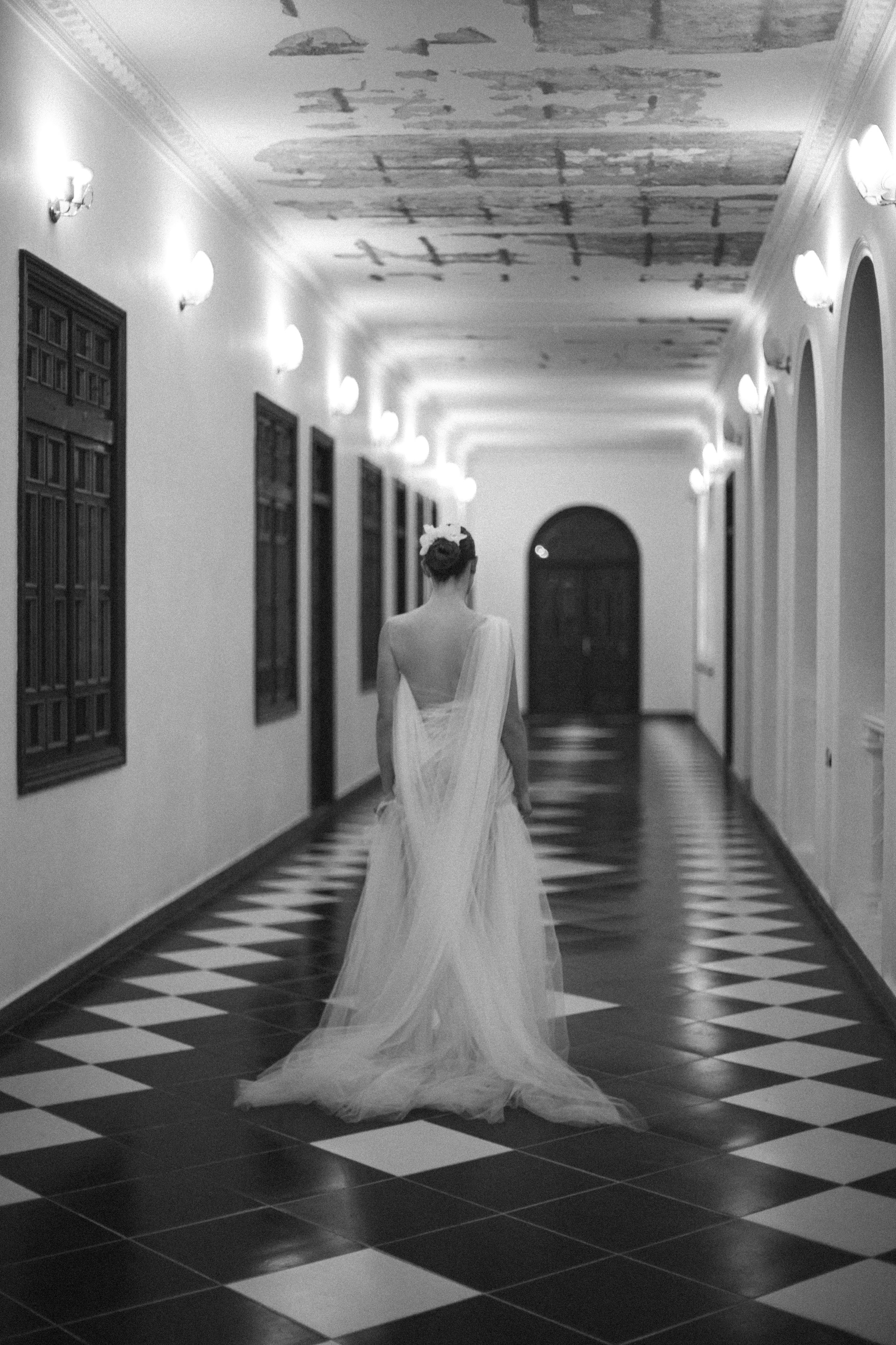 A woman dressed in a wedding gown walking down a corridor with checkered floor tiles and illuminated by wall sconces.