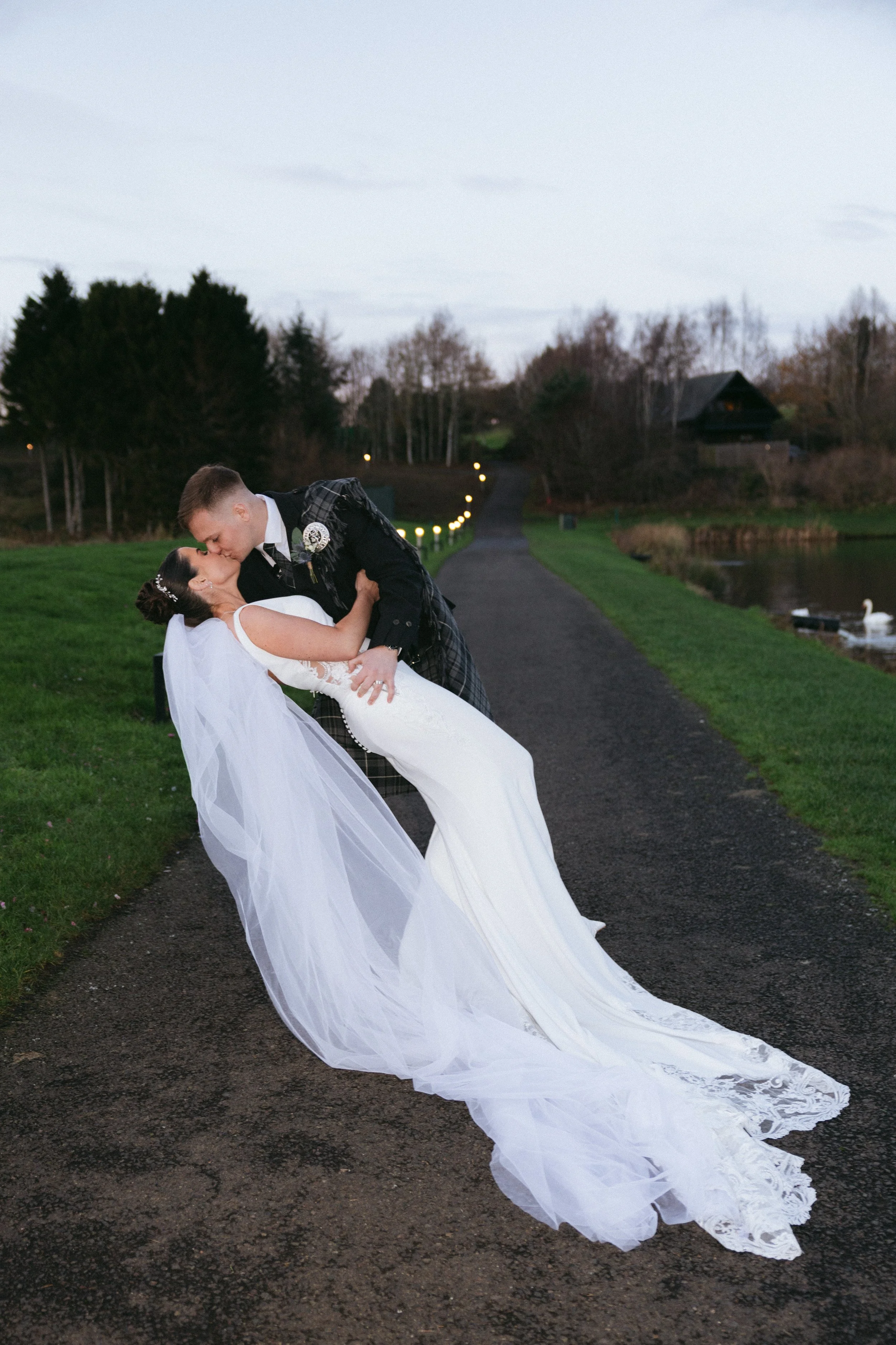 A bride and groom dancing outdoors on a rainy evening with a lake and trees in the background.