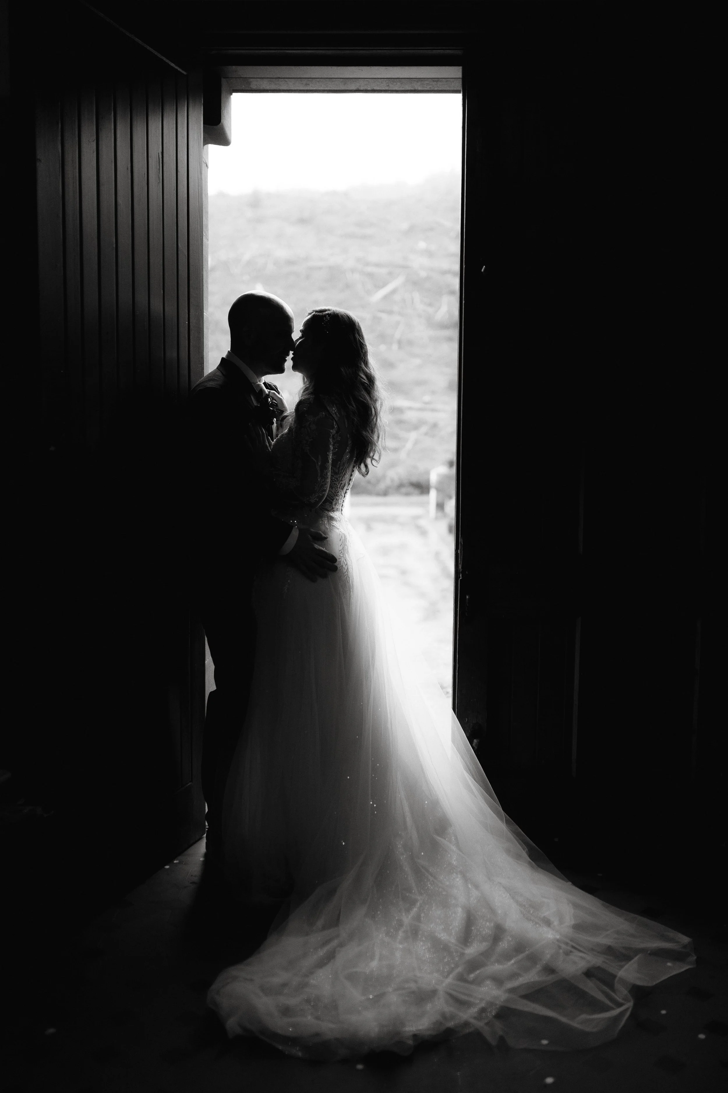 A black and white photo of a bride and groom standing close inside a dimly lit barn or rustic building, about to kiss, with sunlight coming through the open doorway behind them, revealing a blurred outdoor landscape.