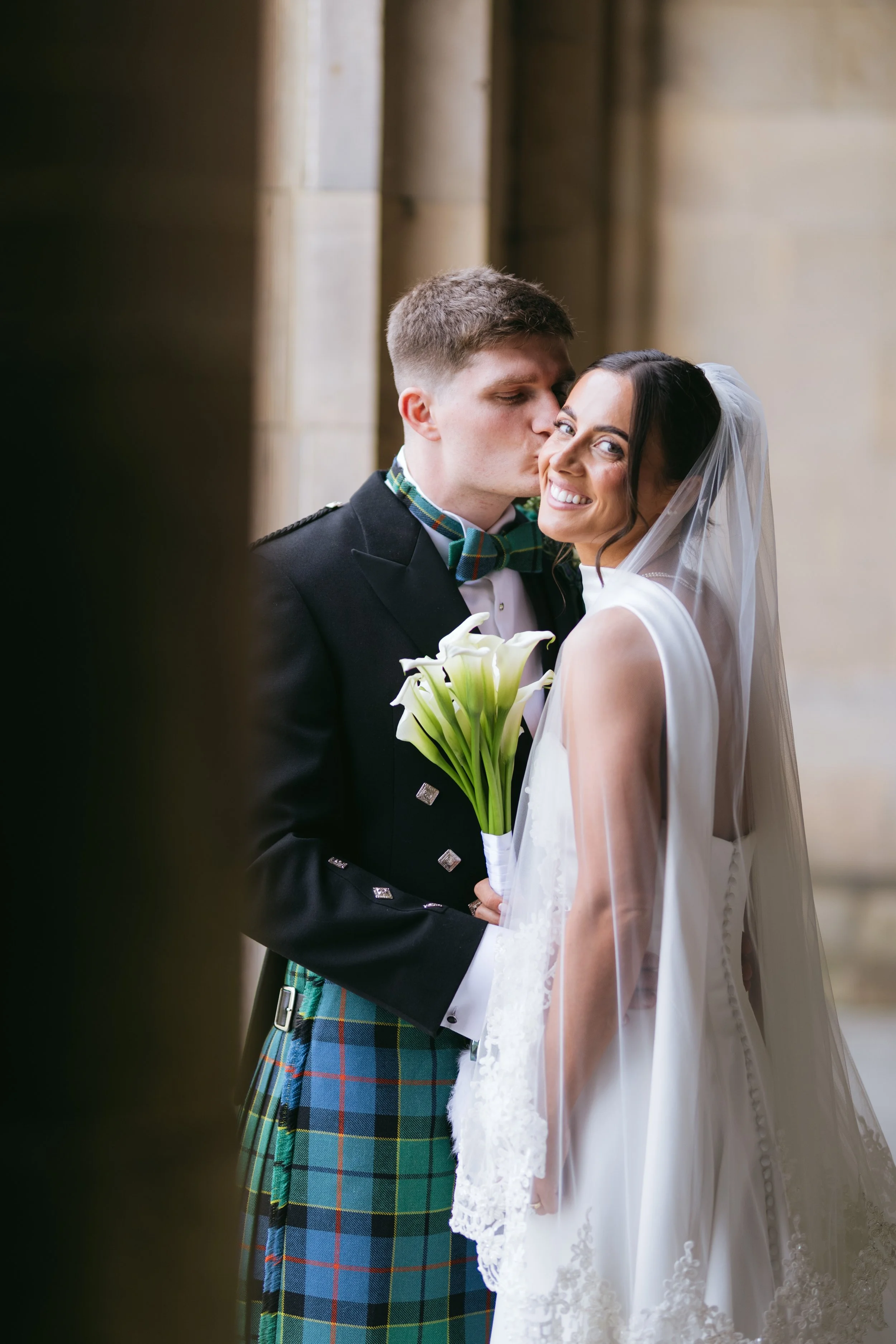 A bride with dark hair wearing a white veil and dress, smiling as a groom in a black suit and green tartan kilt kisses her forehead in a church setting.