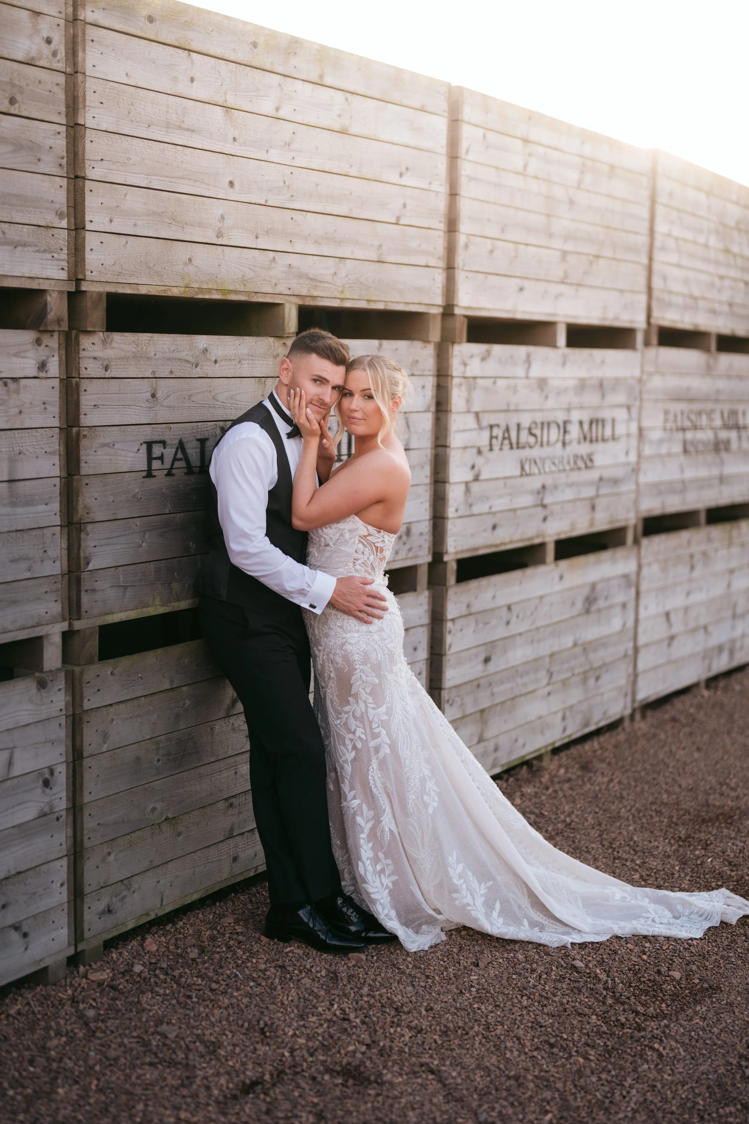 A bride and groom stand close together, posing against a wooden fence at sunset. The bride wears a white lace wedding gown with a train, and the groom wears a black suit with a white shirt and black bow tie. The couple looks directly at the camera, with the groom gently holding the bride's waist and the bride touching the groom's face.
