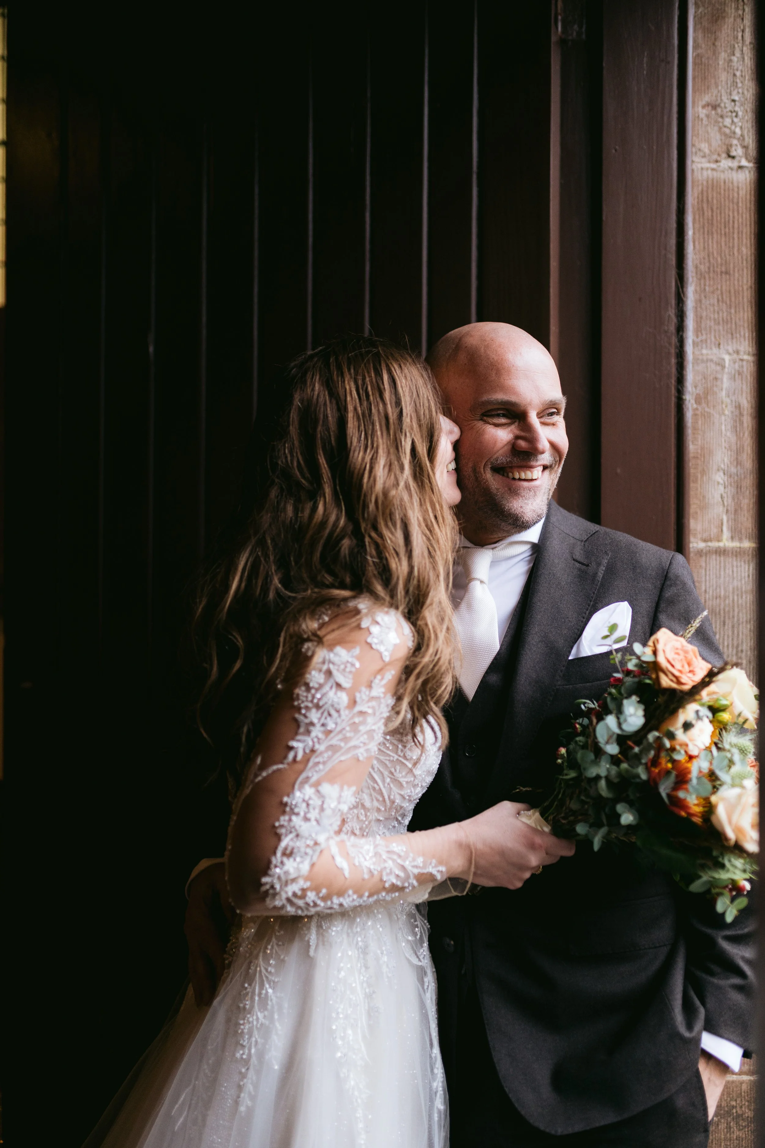 A bride and groom at a wedding, with the bride whispering to the groom and holding a bouquet of flowers, both wearing formal wedding attire, standing against a dark background.