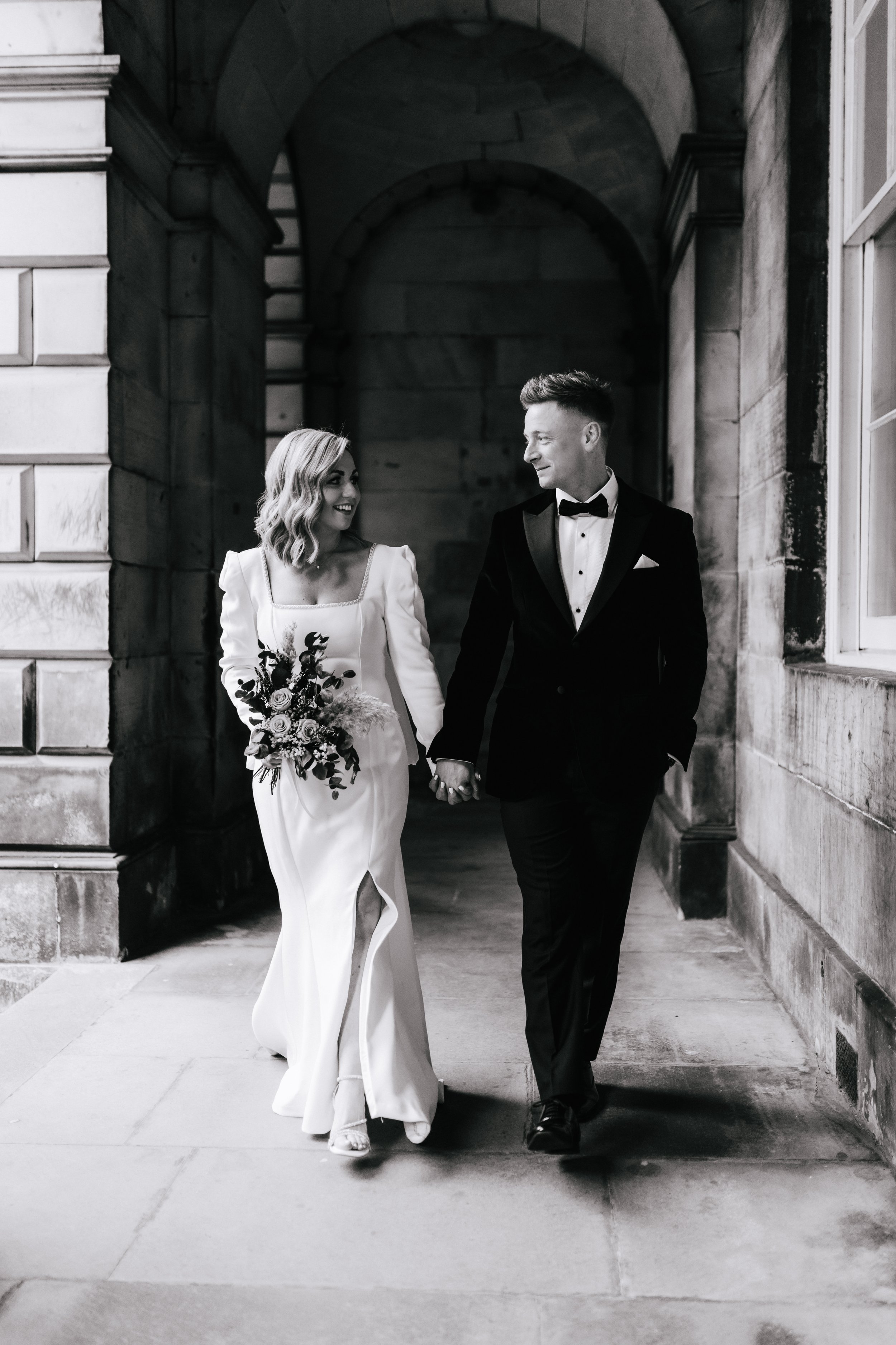 A black and white photo of a bride and groom walking hand in hand. The bride wears a white gown and holds a bouquet, while the groom is dressed in a tuxedo and bow tie. They are smiling and looking at each other, walking under an arched stone structure.