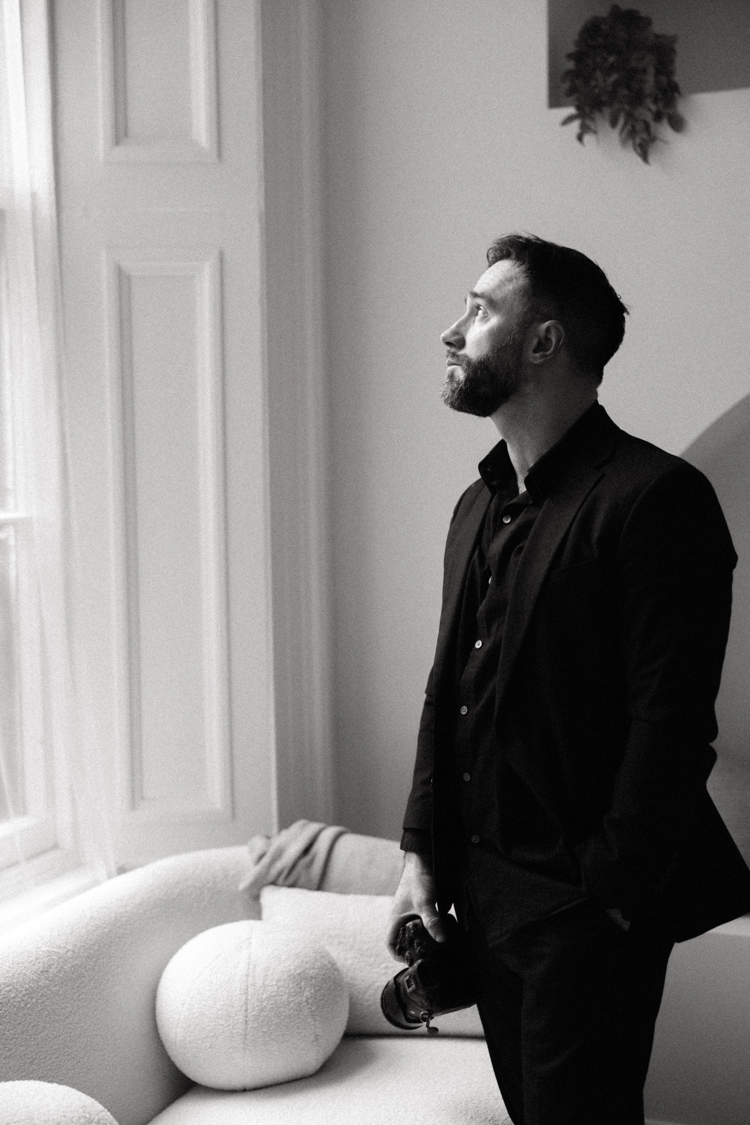 A man with a beard and short hair wearing a dark jacket and black shoes is standing next to a curved, textured sofa with pillows in a room with light-colored walls, a window with white shutters, and a wall hanging of a plant. The photo is in black and white.