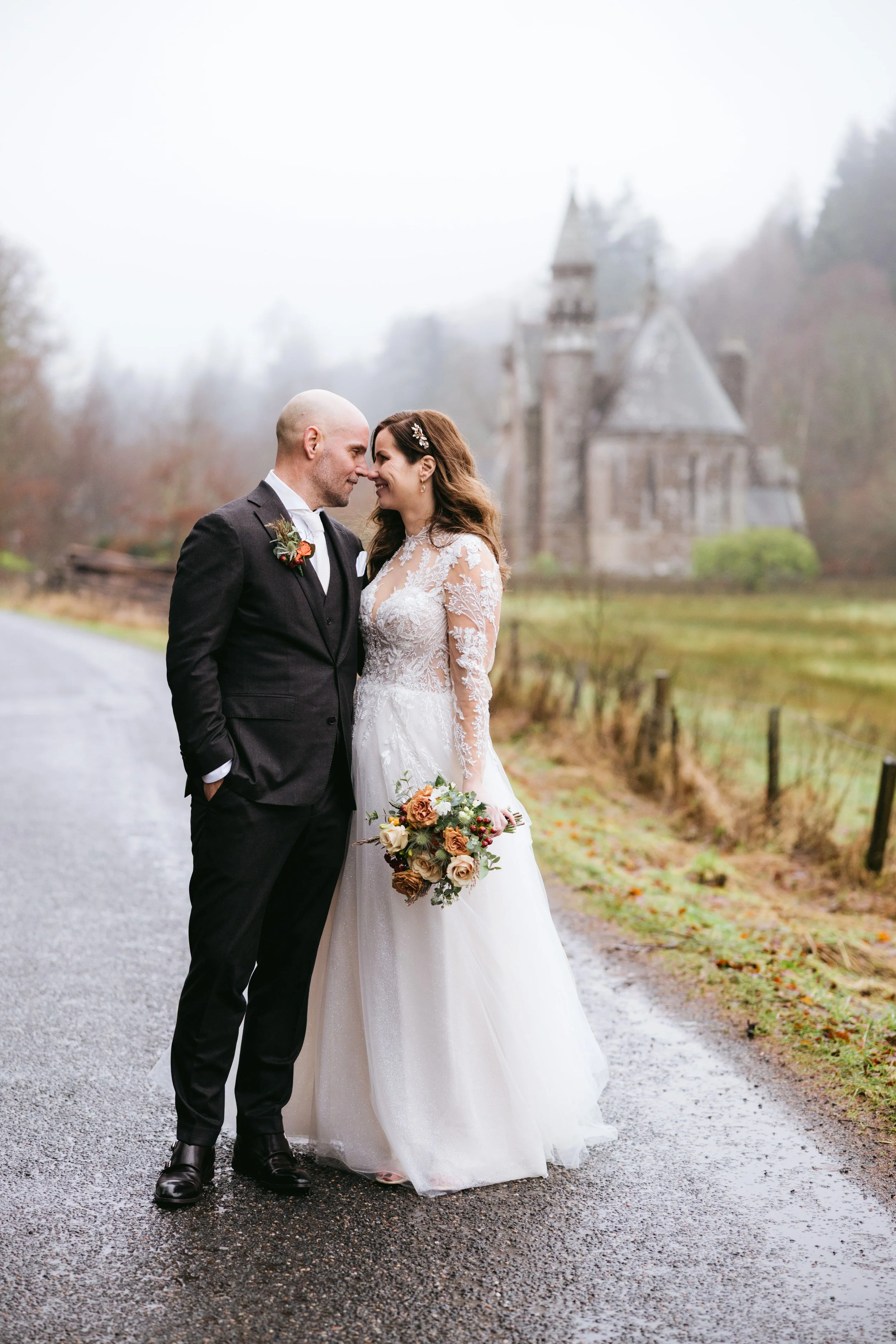 A bride and groom standing close together, touching foreheads, smiling outdoors on a gray, rainy day. The groom is bald, wearing a dark suit with a boutonniere, and the bride has long wavy brown hair with a hairpin, wearing a lace wedding dress, holding a bouquet of flowers. In the background, there is a historic castle, trees, and a wet gravel path.