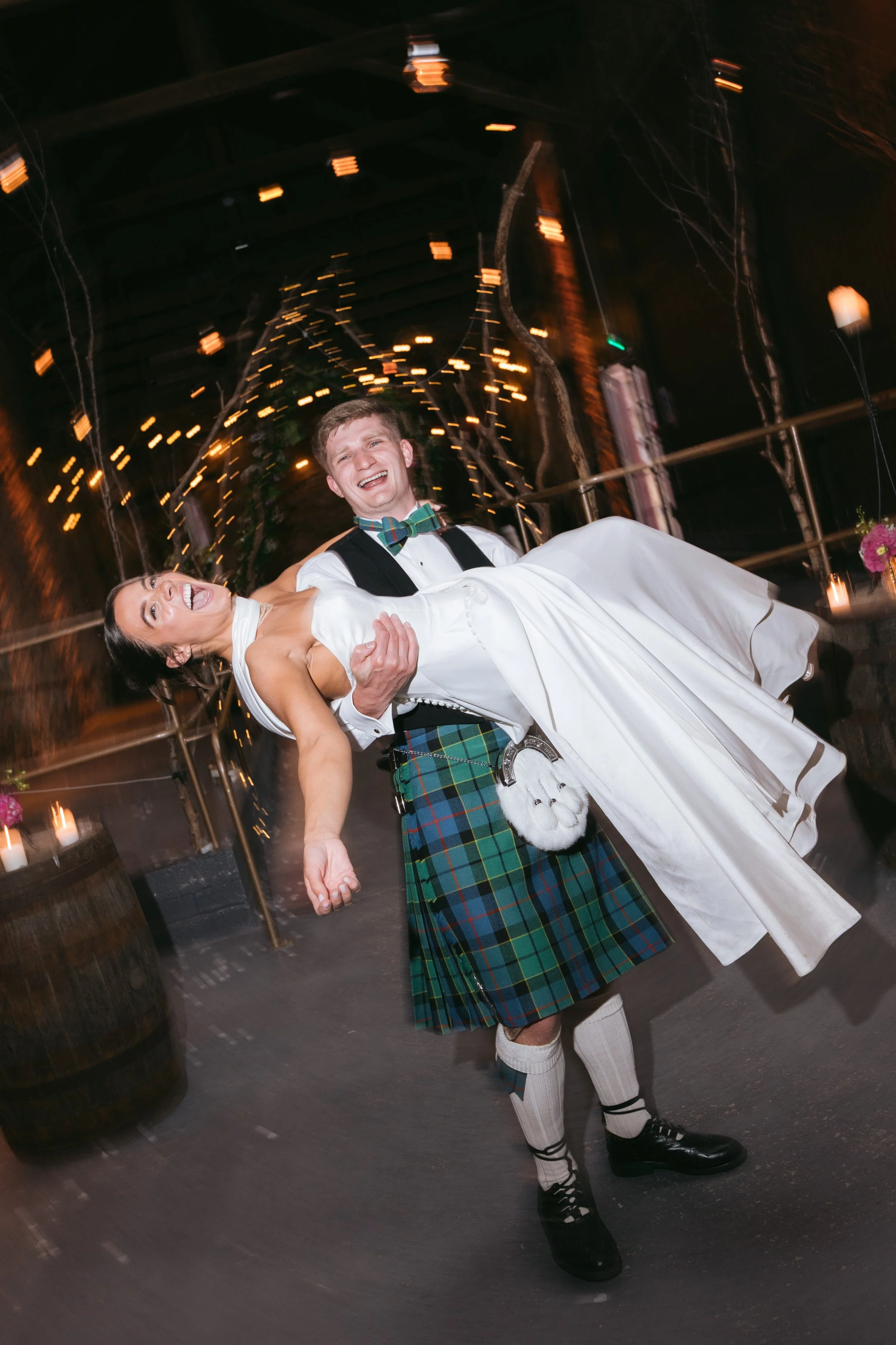 A bride and groom are dancing at their wedding celebration. The groom is wearing a traditional Scottish kilt and the bride is in a white wedding dress, both smiling joyfully. The background has warm lighting and string lights creating a festive atmosphere.