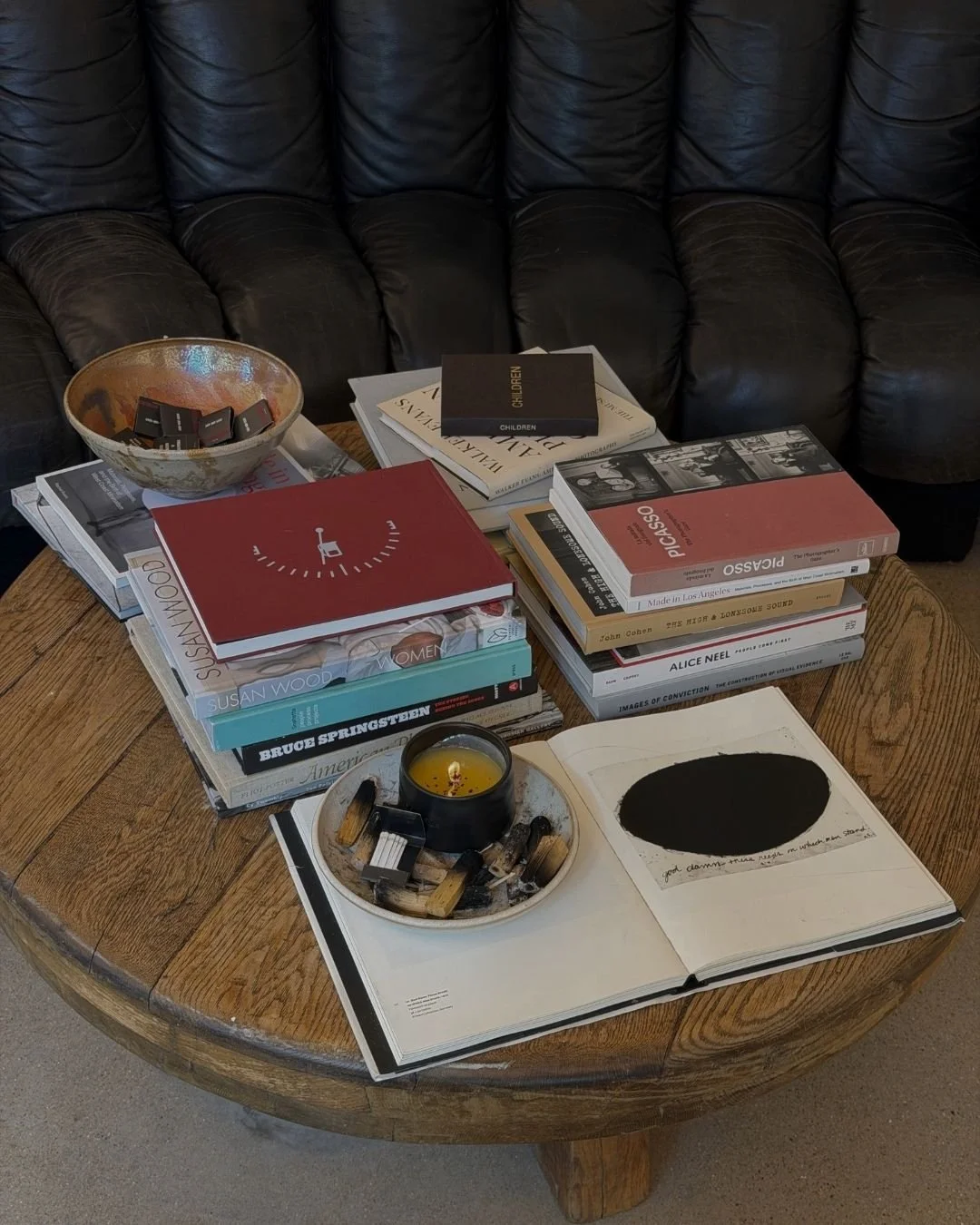 A round wooden coffee table with a black leather couch behind it. The table has a stack of art and photography books, a black candle in a bowl, and an open book with a black oval on one page and text on the opposite page. There is also a ceramic bowl with small objects inside.