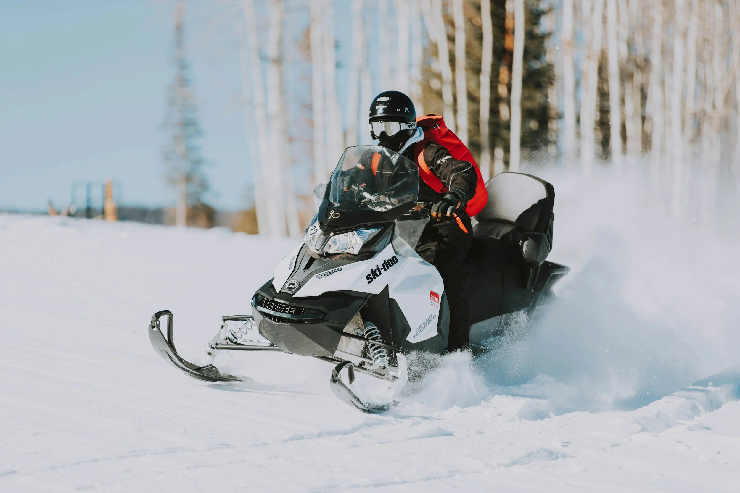 Person riding a snowmobile on snow-covered terrain, wearing a black helmet, goggles, black jacket, red backpack, and gloves, with a forest of leafless trees in the background.