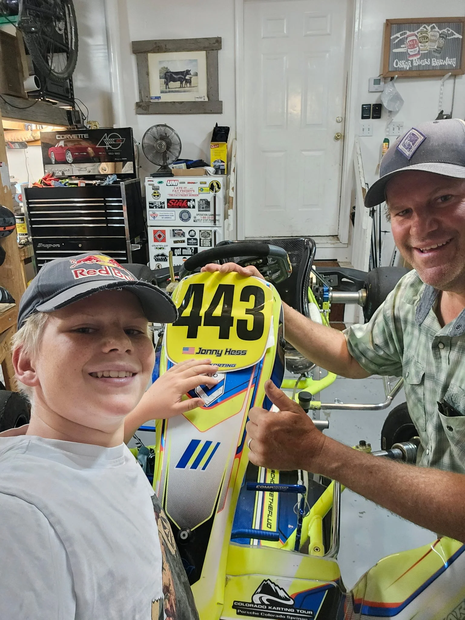Two males smiling, one child and one adult, posing with a yellow go-kart, in a garage or workshop, with tools and stickers in the background.