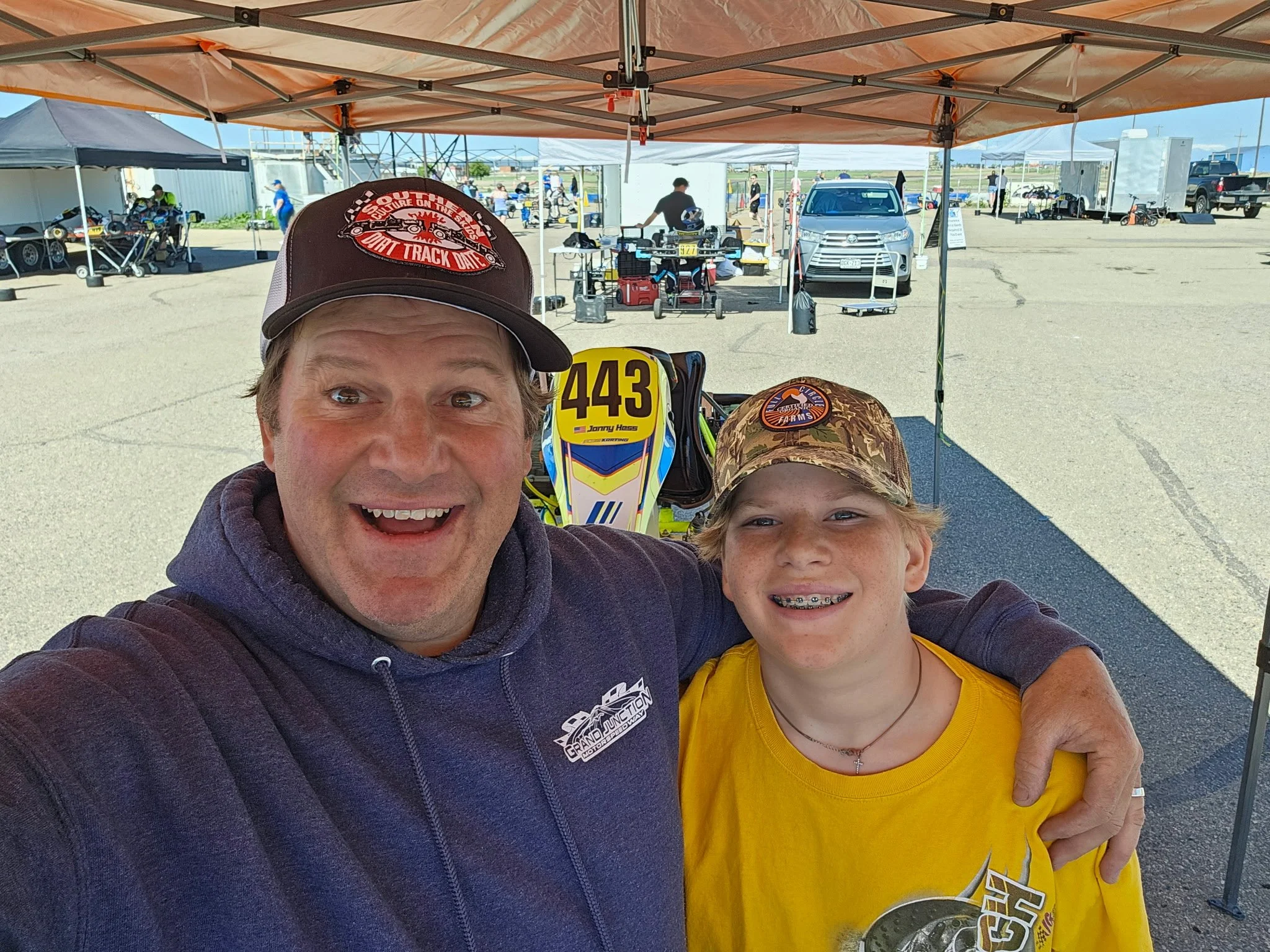 A man and a girl smiling at a go-kart racing event under a canopy tent, with various equipment and vehicles in the background.