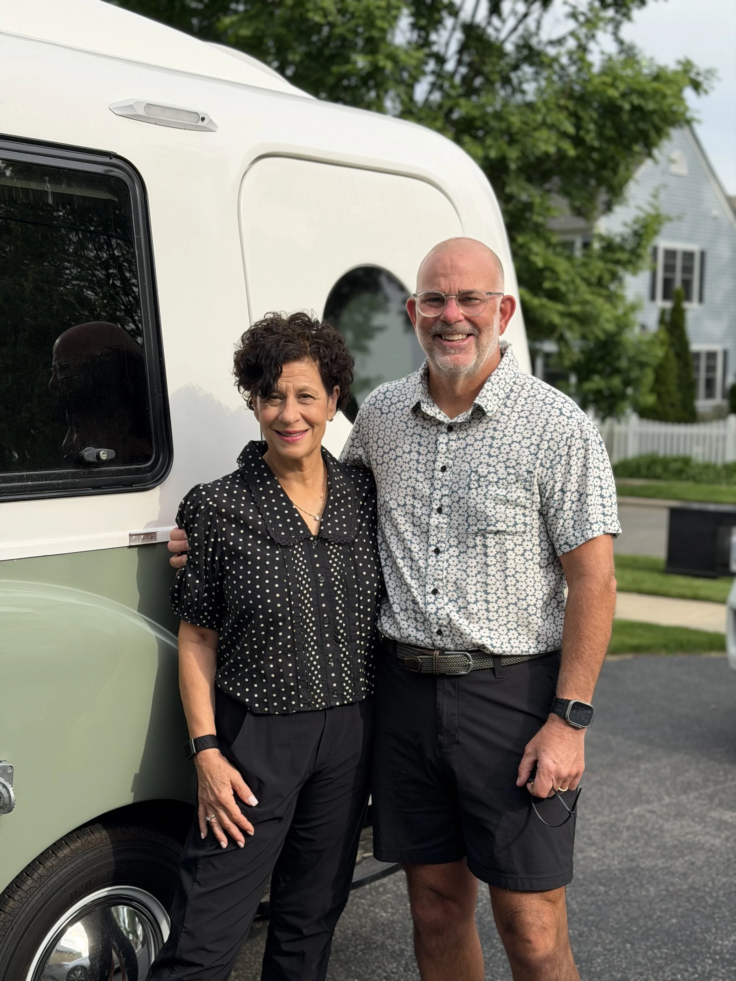 A smiling older woman and a smiling middle-aged man standing outside, near a light green vintage camper van, with green trees and a house in the background.