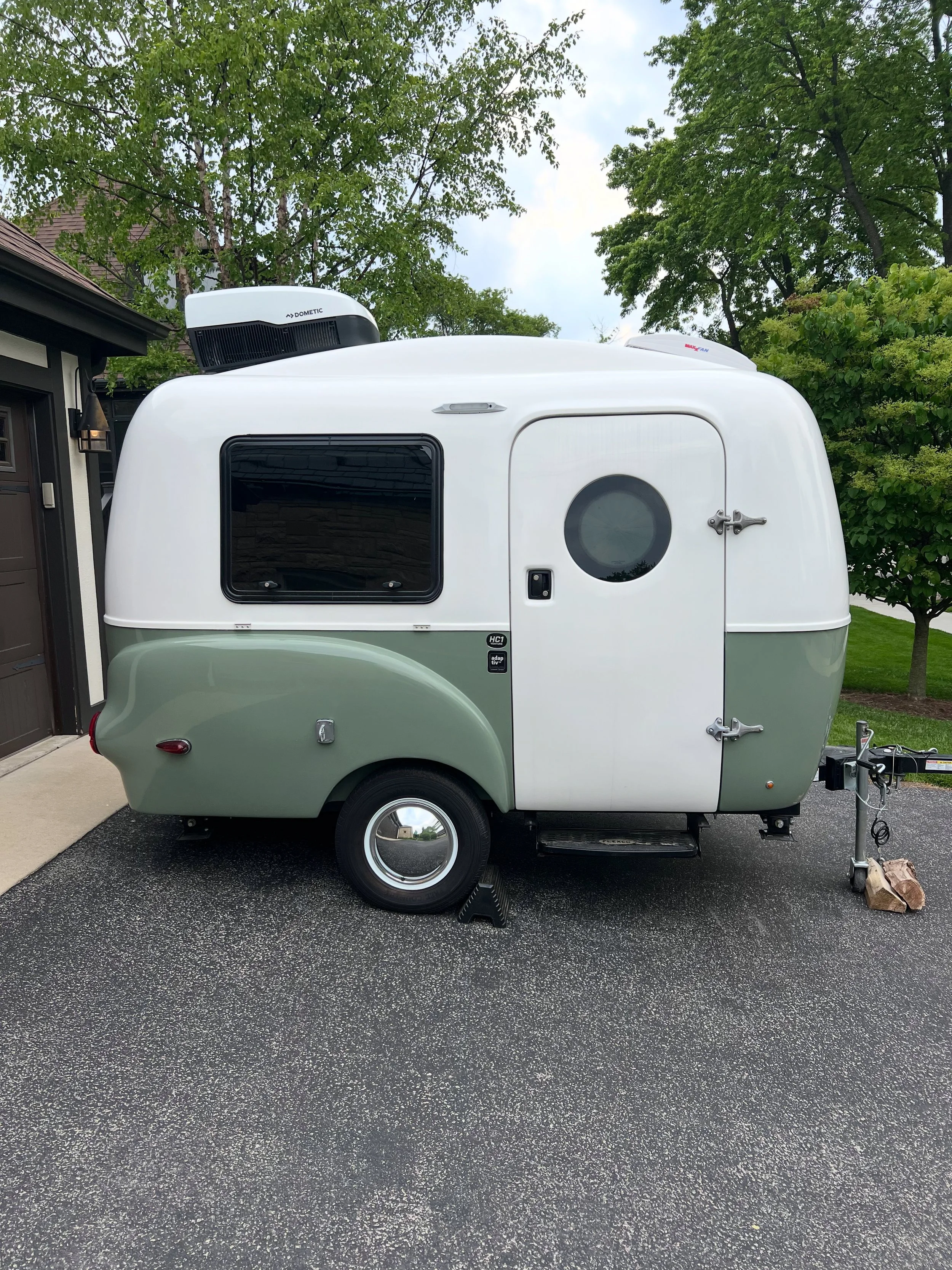 A small travel trailer parked in a driveway with trees and a house in the background.