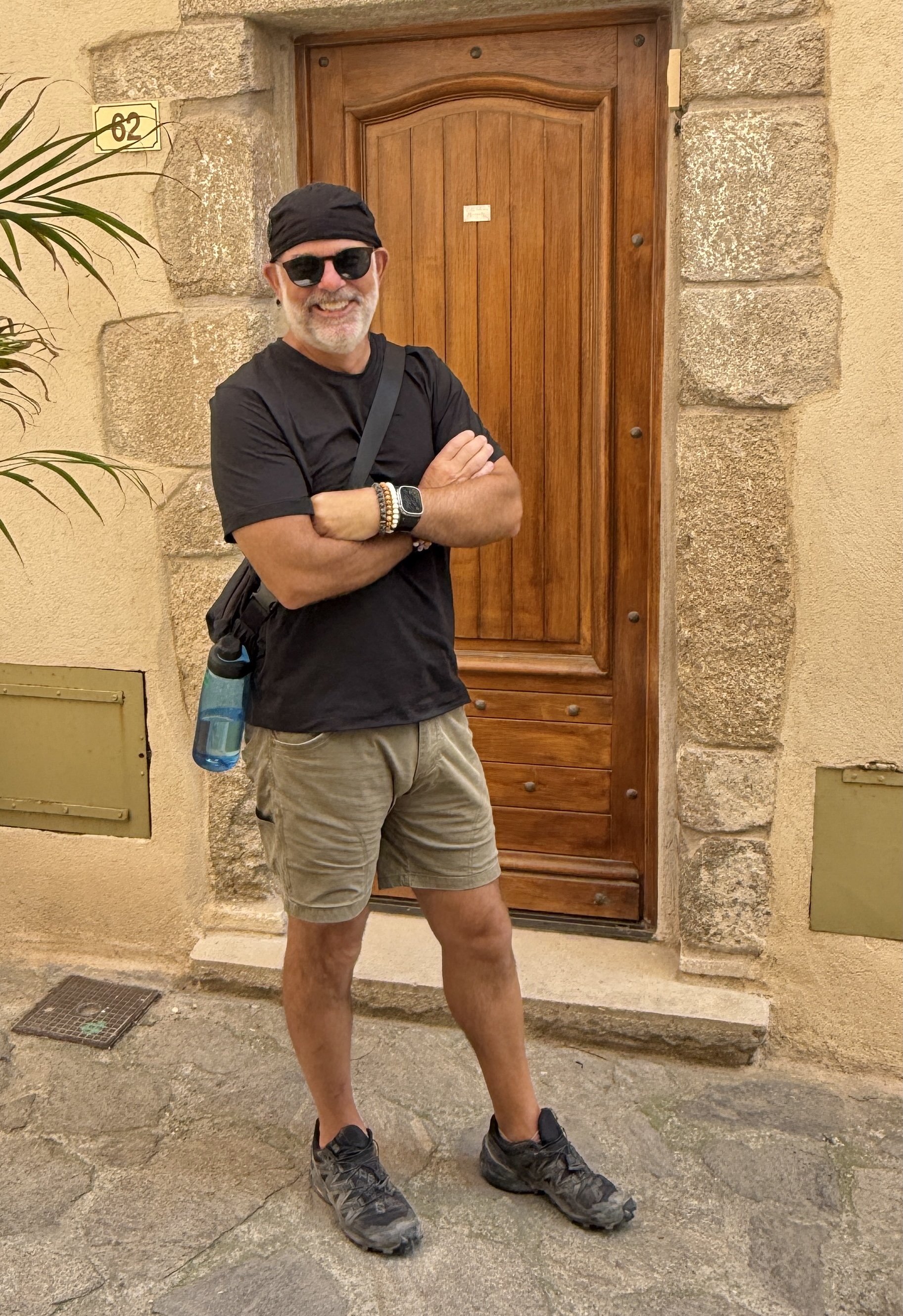 A smiling man, Shaun Emerson, wearin sunglasses, a black headband, a black t-shirt, khaki shorts, and hiking shoes, standing outside in front of a wooden door with stone accents, crossing his arms, with a water bottle hanging from his backpack strap.