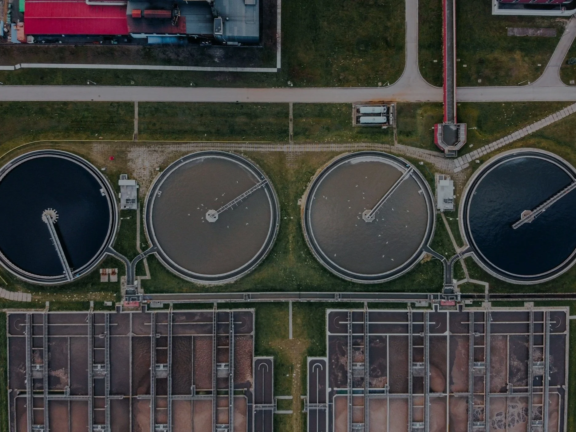 Aerial view of a waste water treatment facility with three large circular clarifiers filled with water, connected by walkways and surrounded by grass, roads, and buildings.