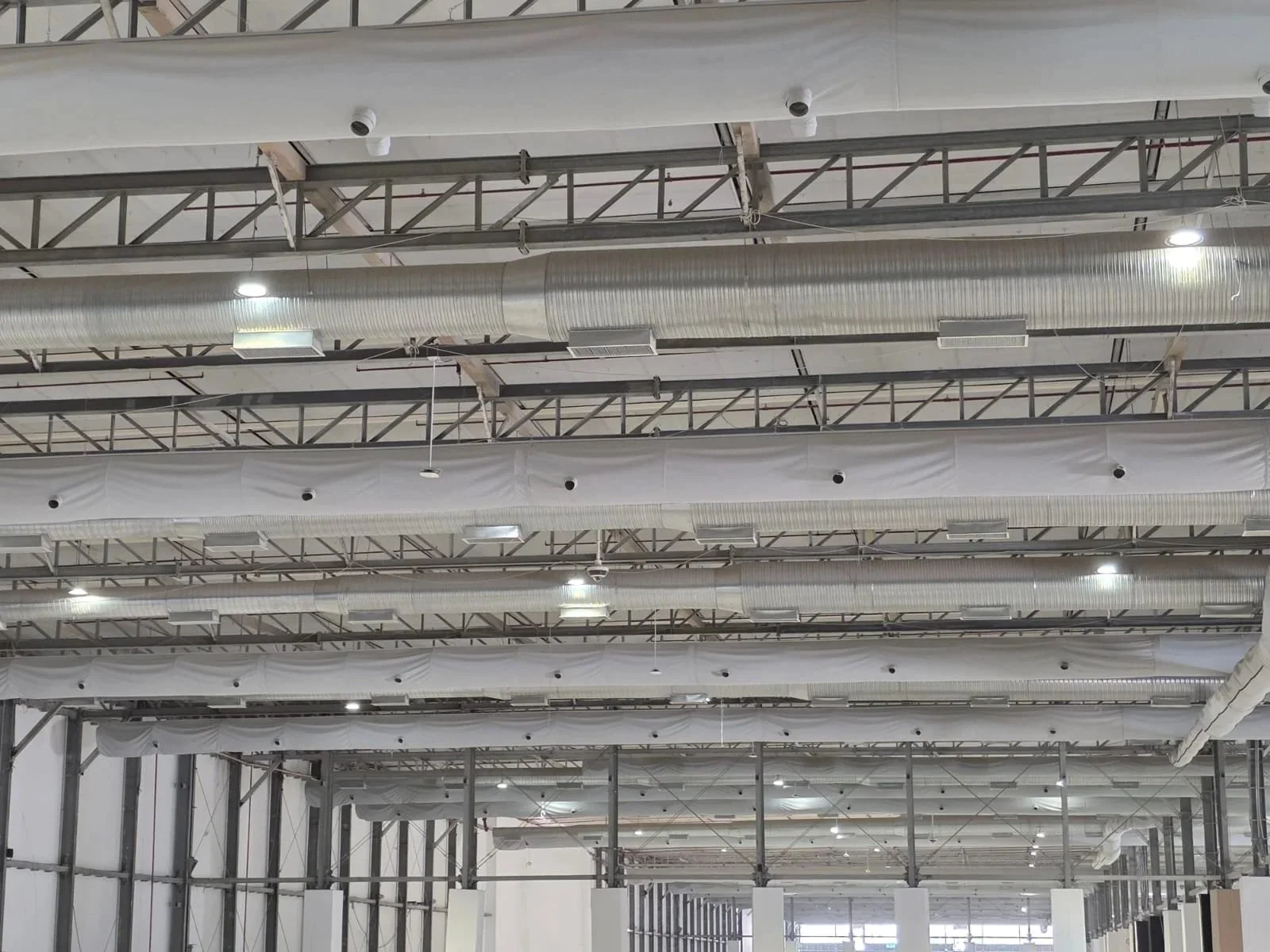Interior of a large industrial or commercial building with exposed metal framework, large ductwork, and grey fabric ducts hanging from the ceiling.
