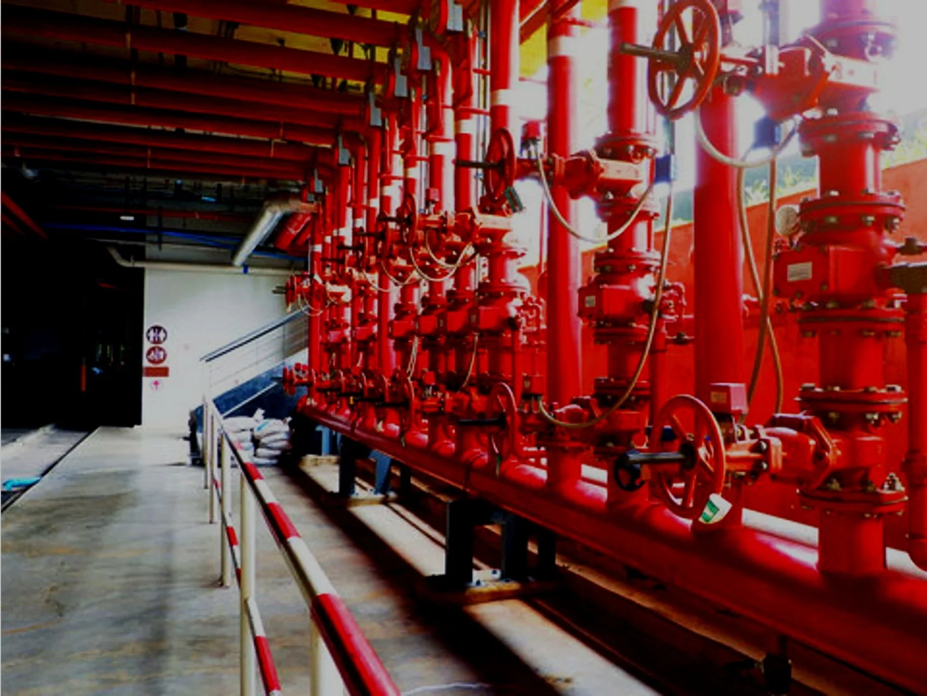 Inside a large industrial facility with a row of red fire protection pipes and valves, safety barriers, and a conveyor belt on the left side.