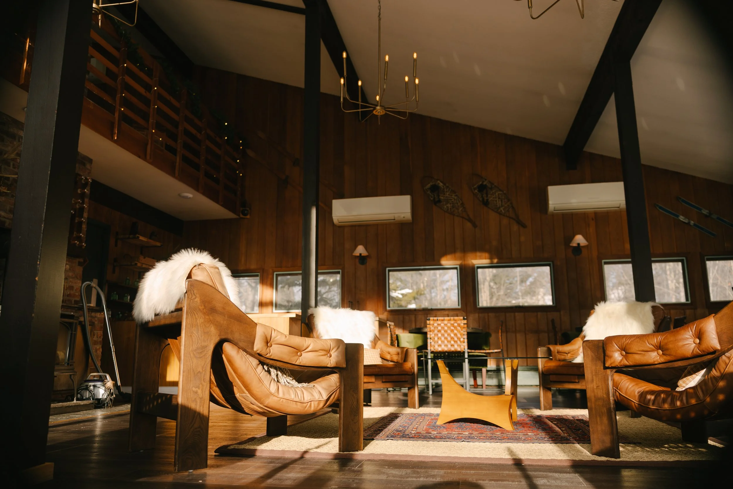 Cozy living room with wooden walls, brown leather armchairs with white fur throws, a colorful area rug, and large windows letting in natural light. Decor includes vintage snowshoes and skis on the wall, two air conditioning units, and a modern chande
