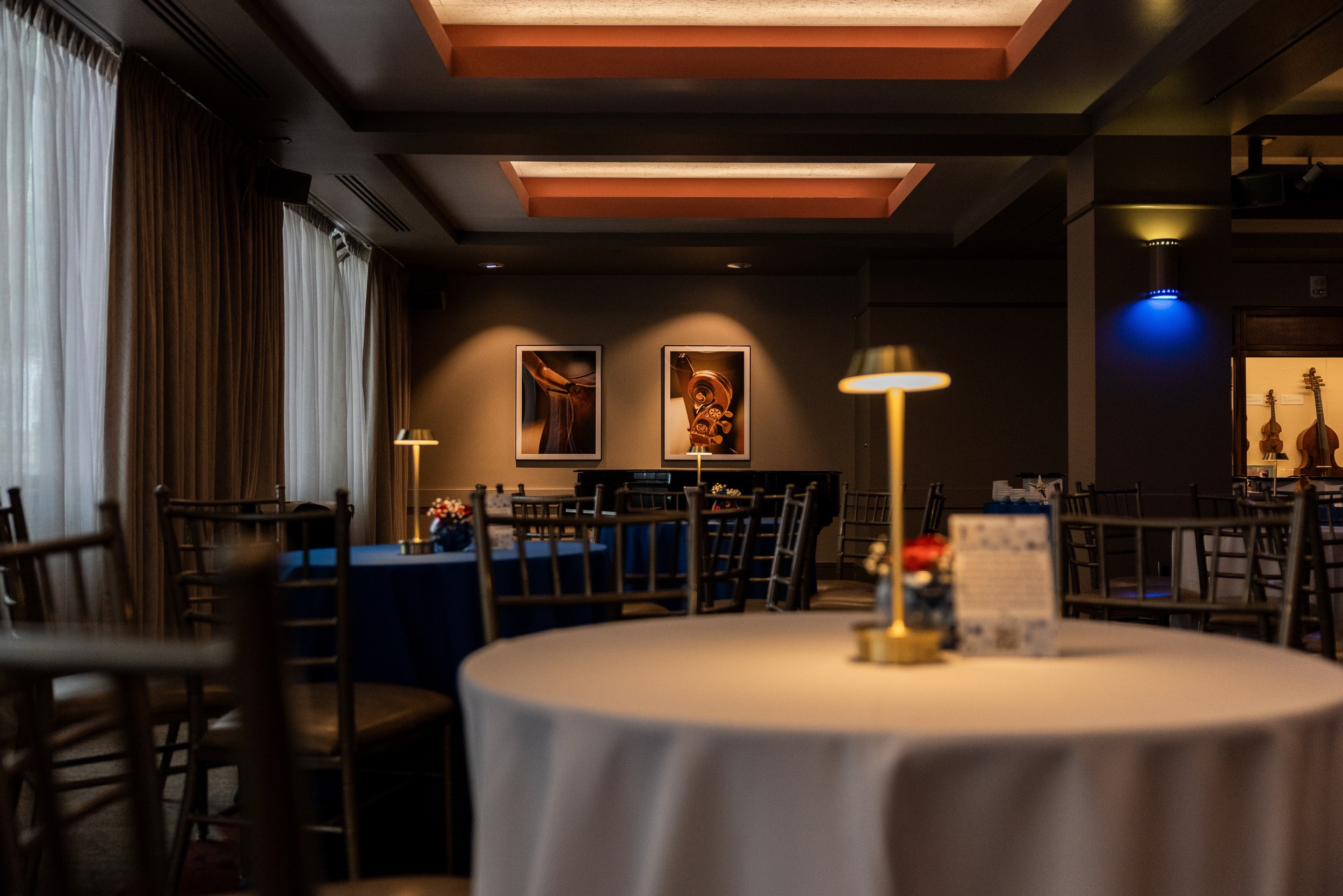 Elegant dining area with round tables covered in white tablecloths, surrounded by wooden chairs, soft lighting, and wall art of musical instruments, with curtains and a ceiling with recessed lighting.