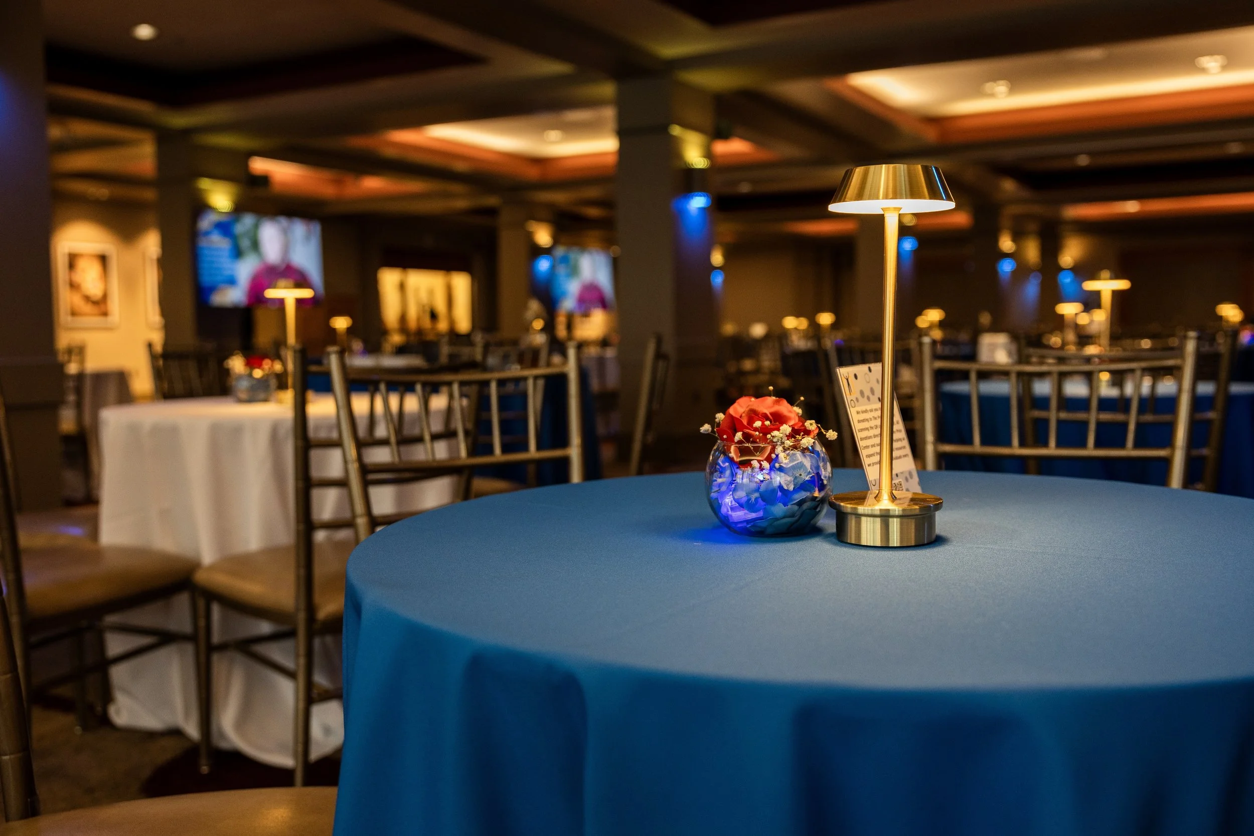 Elegant dinner event in a dimly lit banquet hall with a round table covered with a blue tablecloth, featuring a gold lamp and a small flower arrangement in a glass vase.