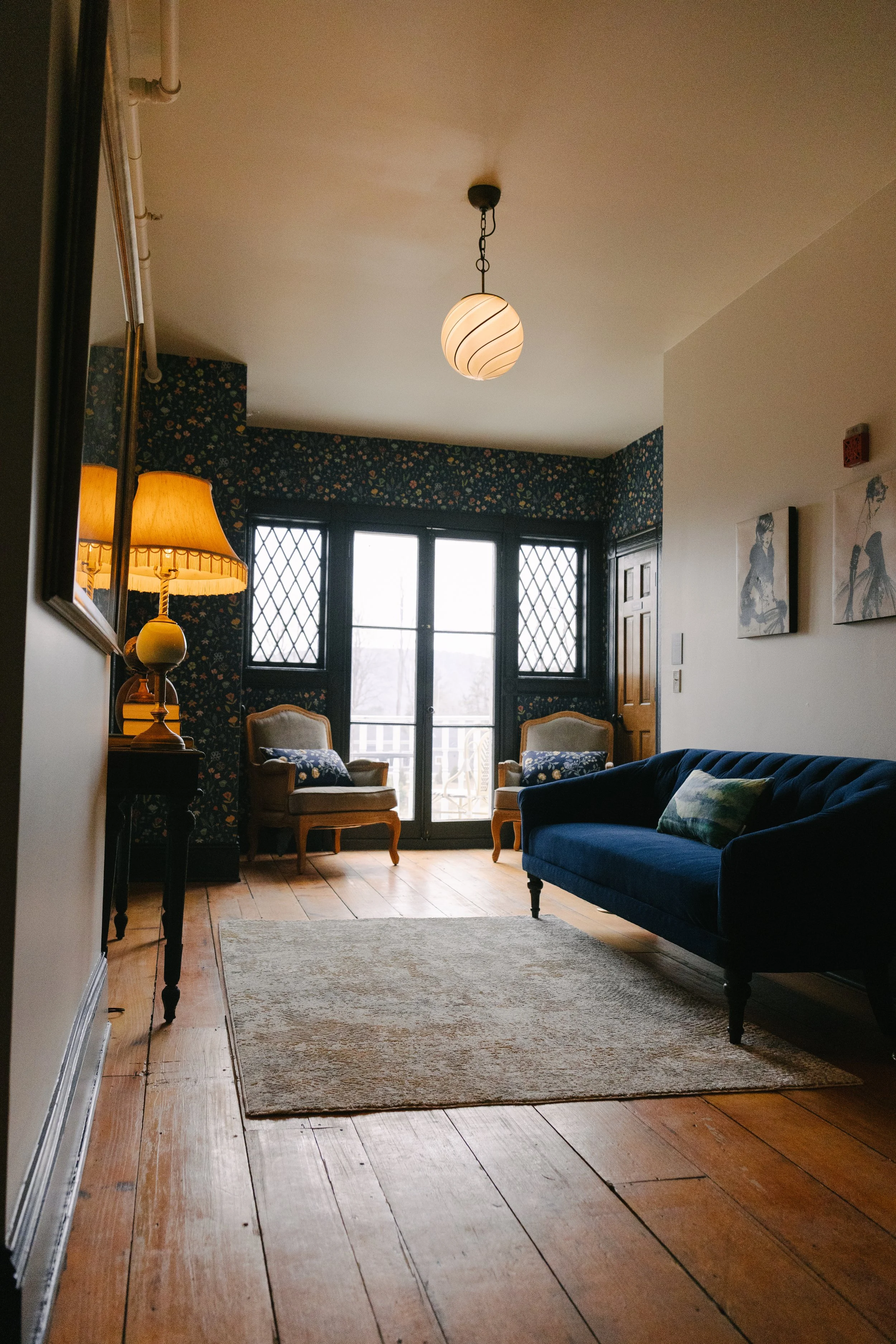 Living room with hardwood floor, a blue sofa, two beige armchairs, a table lamp with a yellow shade, a hanging ceiling light, and a pair of glass doors leading outside.