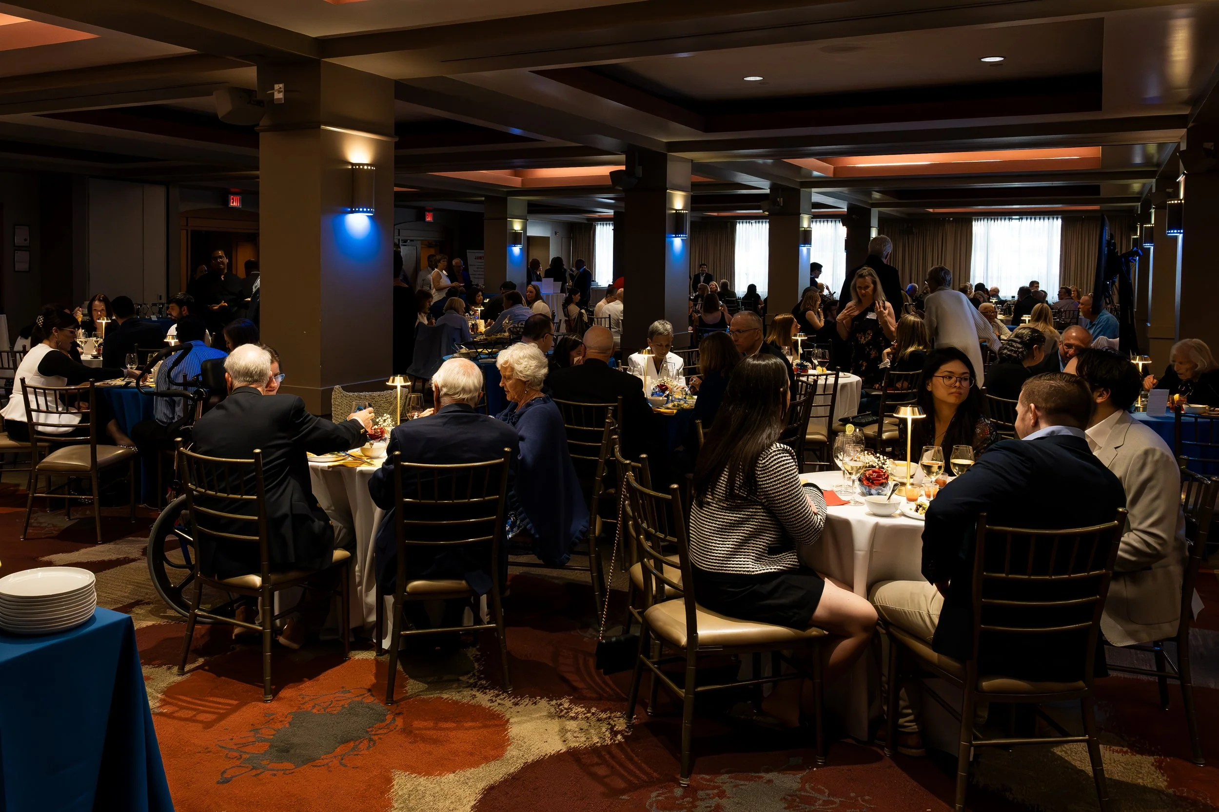 A large banquet hall filled with people seated at tables, dining and talking. The room has soft lighting with blue accent lights on the columns and a carpet with a red and beige pattern. Some attendees are visible in the background, and a few servers