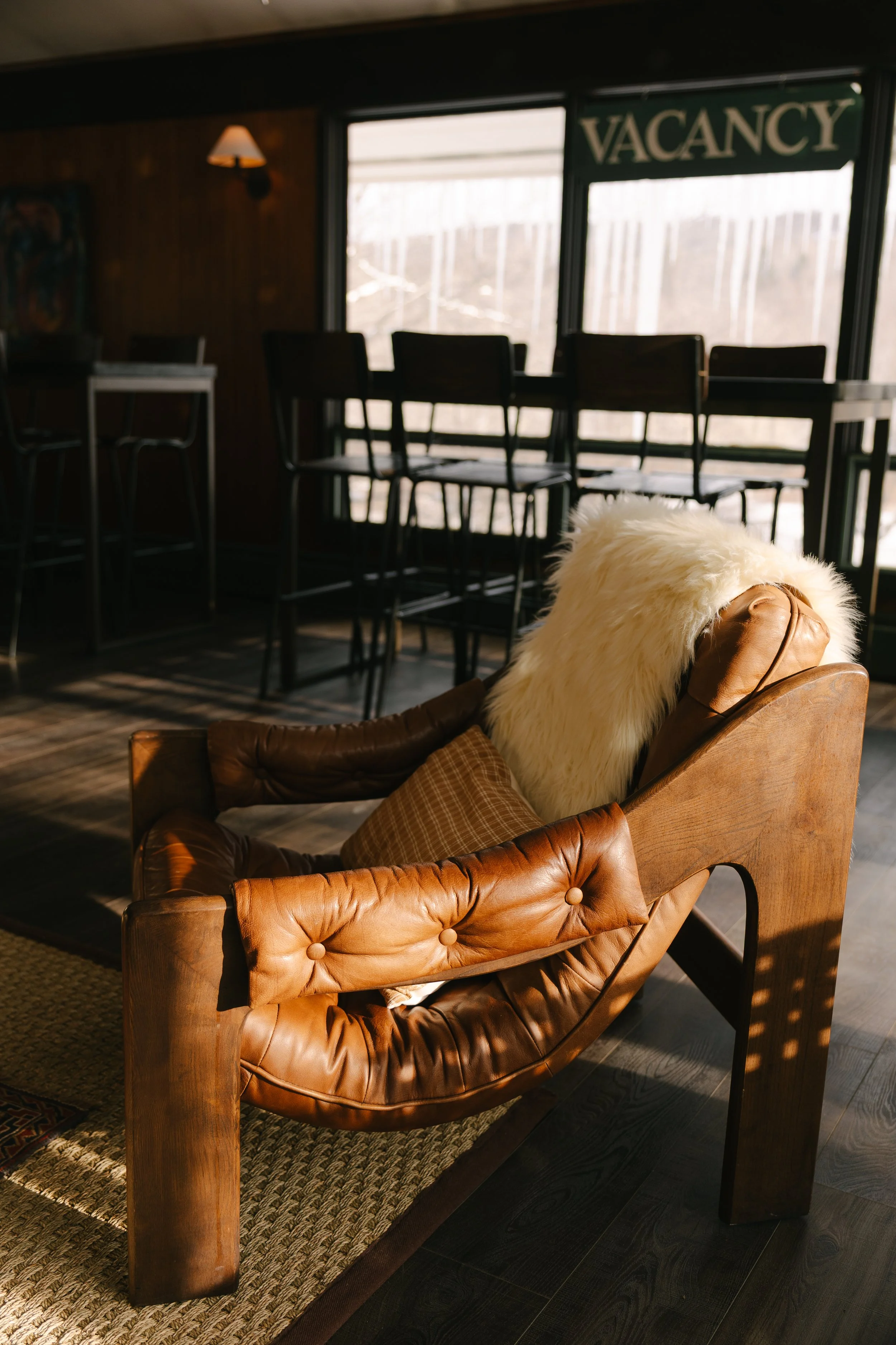 Empty bar or restaurant interior with a large window, a "VACANCY" sign, and a leather chair with fur and pillow inside.