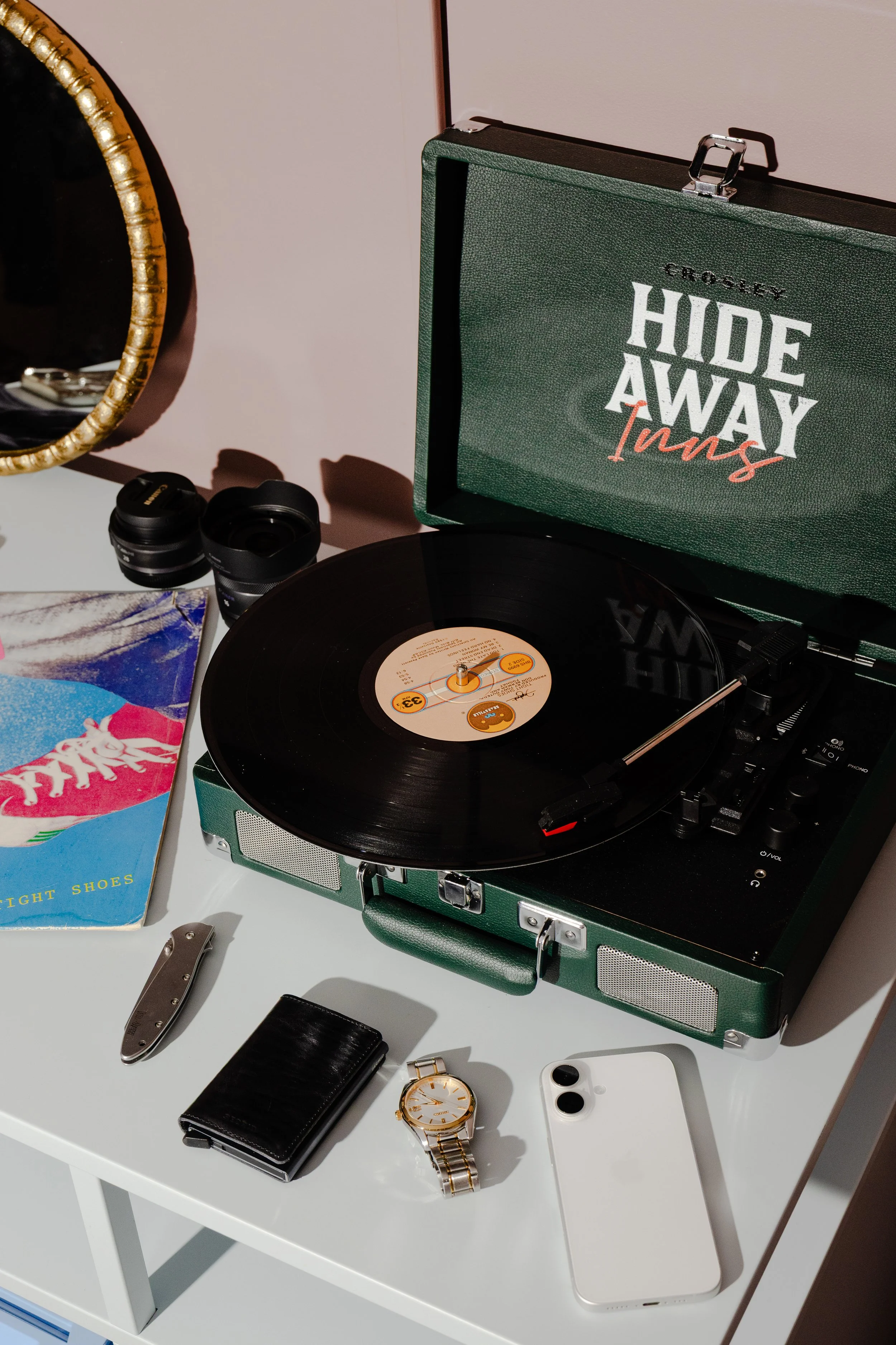 A vintage green portable record player playing a vinyl record. Surrounding it are a smartphone, a black wallet, a gold wristwatch, a small folding knife, two camera lenses, a colorful magazine, and a mirror with a gold frame. The record player has an