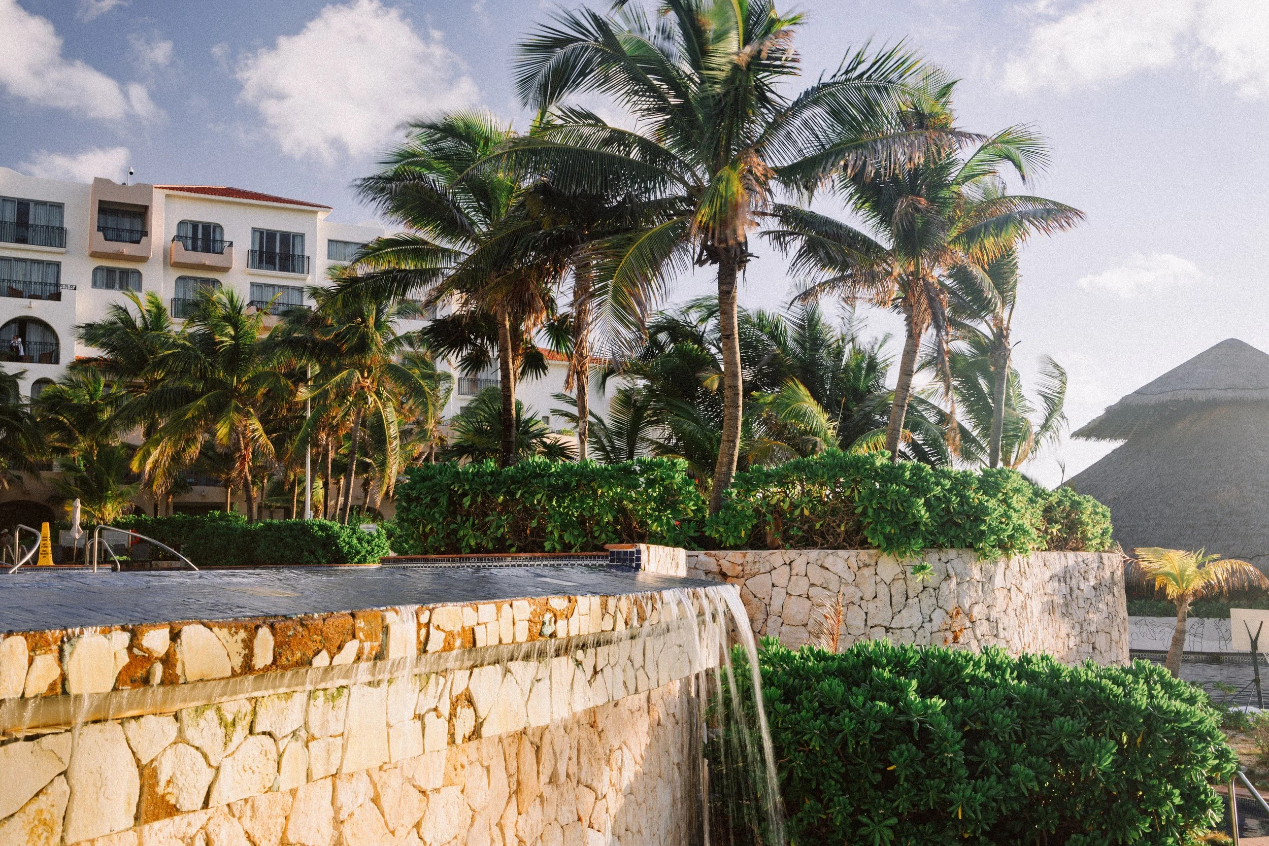Pool with waterfall feature, stone wall, lush green plants, and tall palm trees in a tropical resort setting with a large white building in the background and a thatched roof hut.