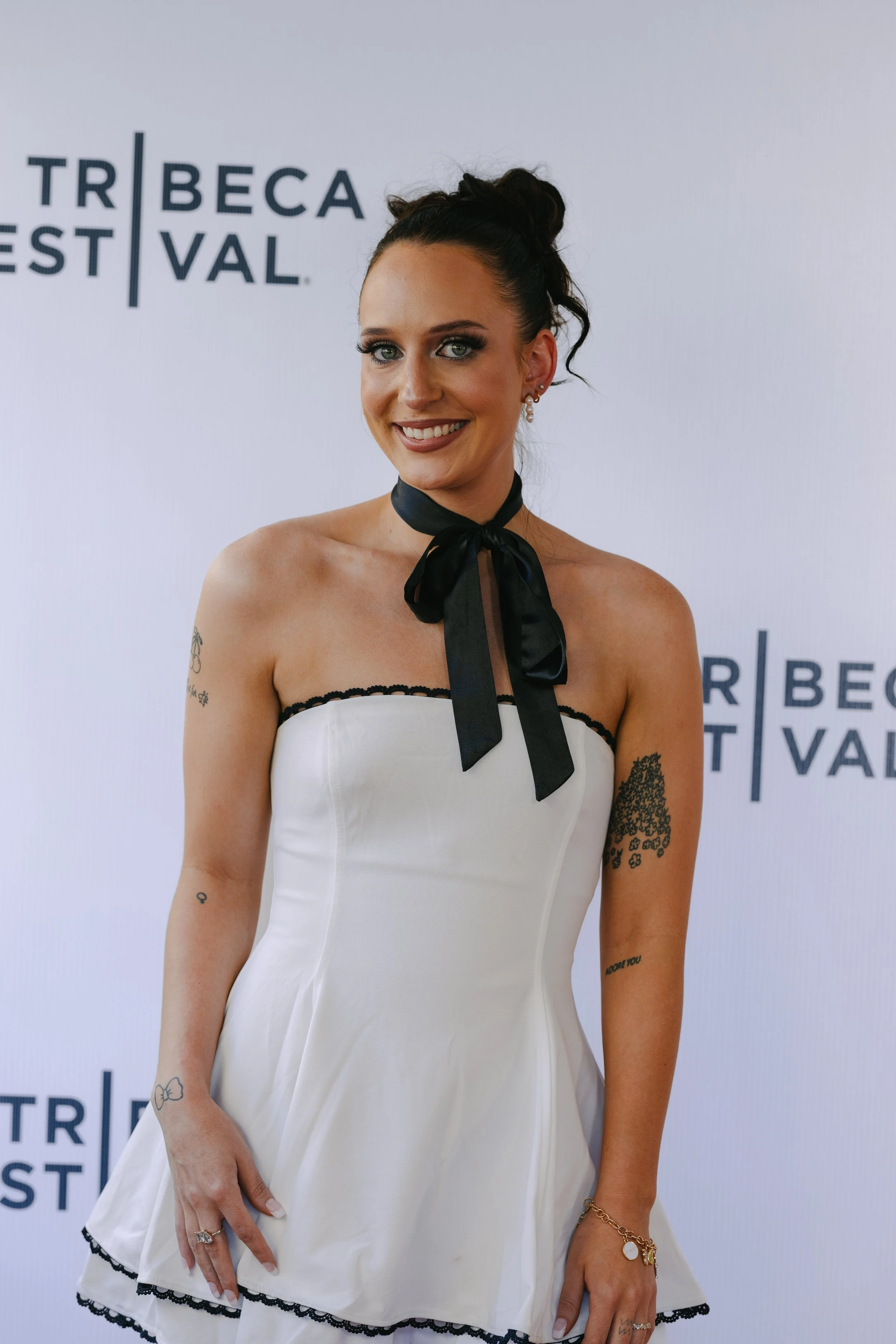 A woman with dark hair styled in an updo wearing a strapless white dress with black trim and a black necktie, smiling at a film festival event, with tattooed arms and jewelry.