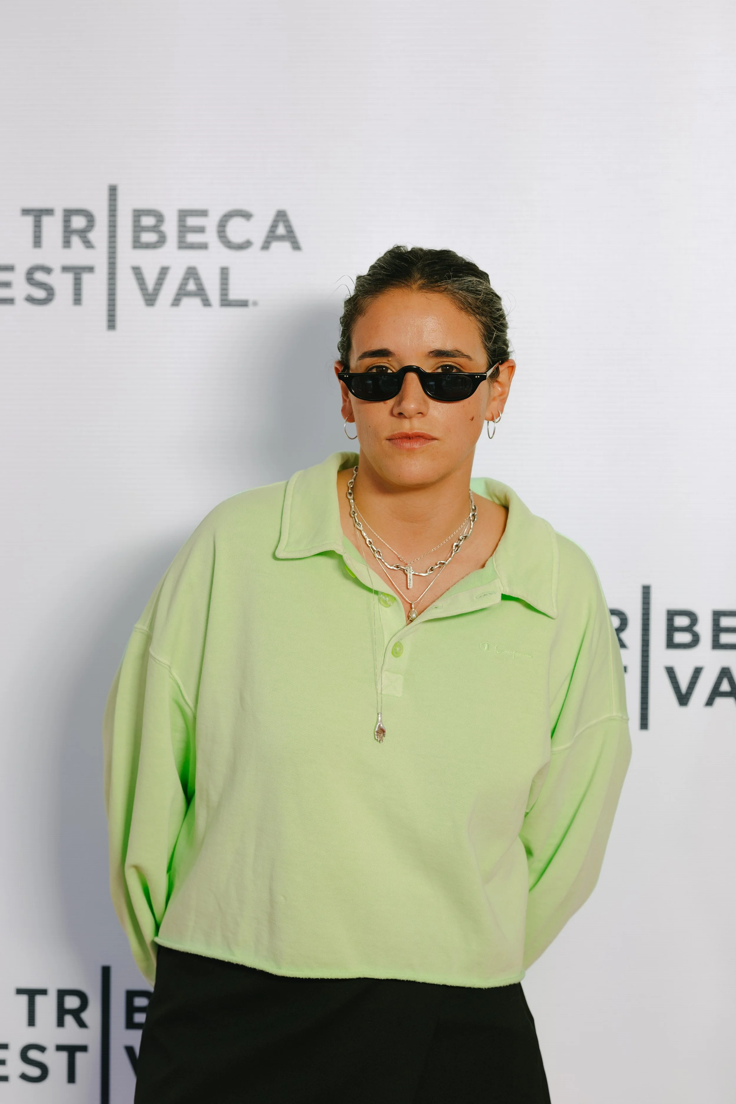 A woman standing in front of a white backdrop with the Tribeca Film Festival logo. She is wearing black sunglasses, a light green oversized polo shirt, and layered silver necklaces, with her hair pulled back.