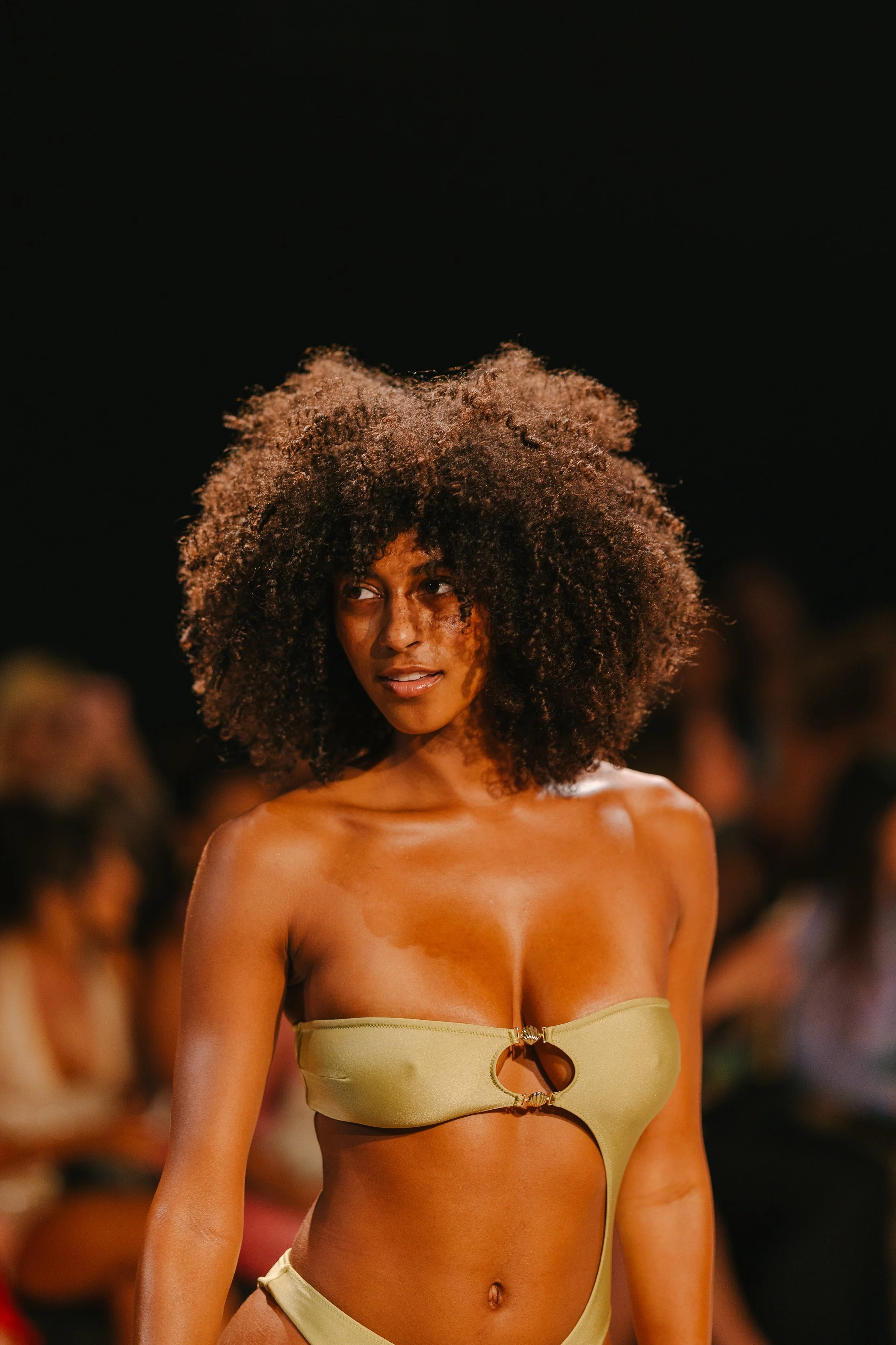 A woman with curly hair walking on a runway wearing a light green bikini with a ring detail in the center.