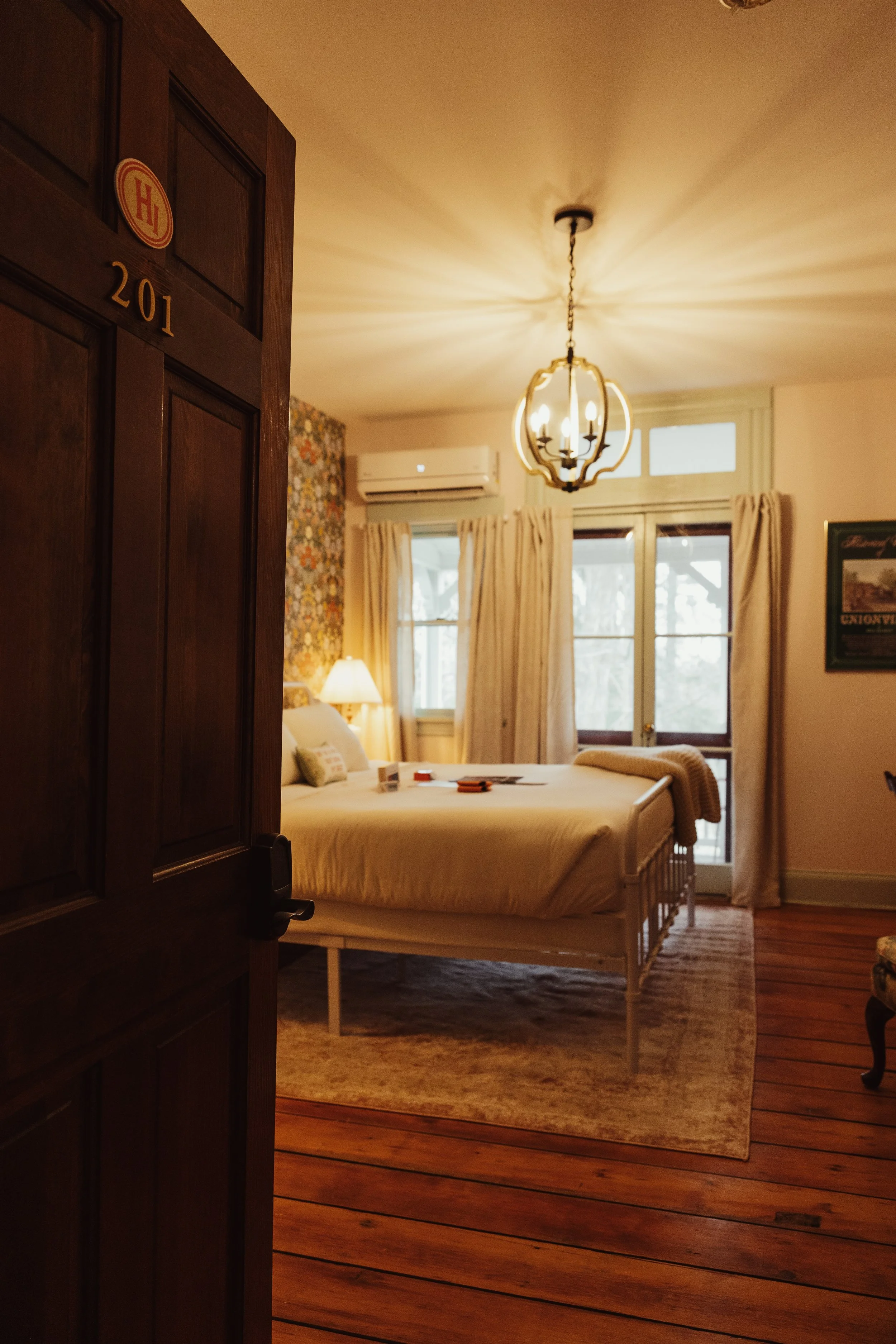 View of a cozy bedroom with a bed, side lamp, large windows with cream curtains, wooden floor, and vintage chandelier lighting.
