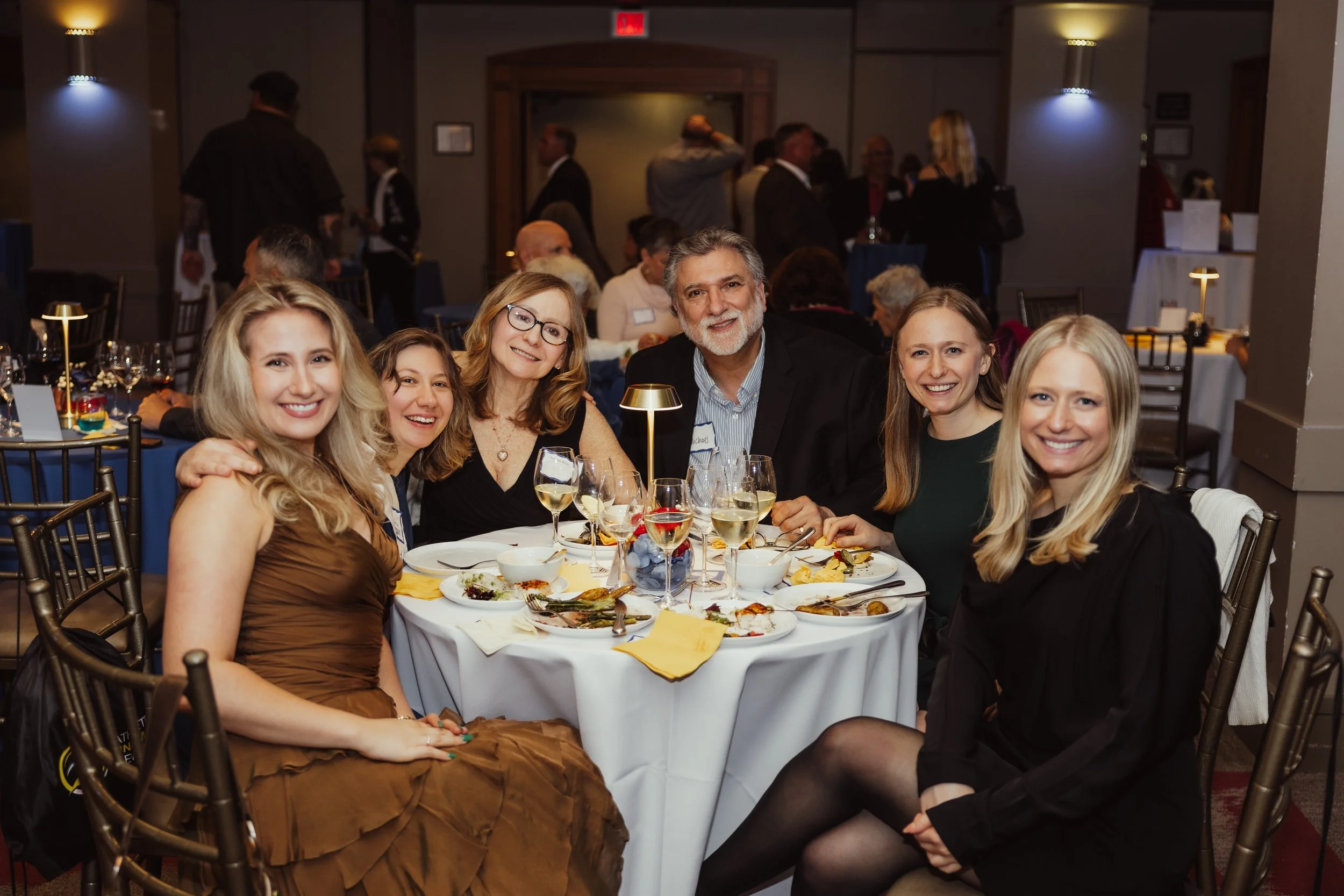 A group of five people, three women and two men, sitting around a dining table at a formal event, smiling at the camera, with plates of food and glasses of wine in front of them.
