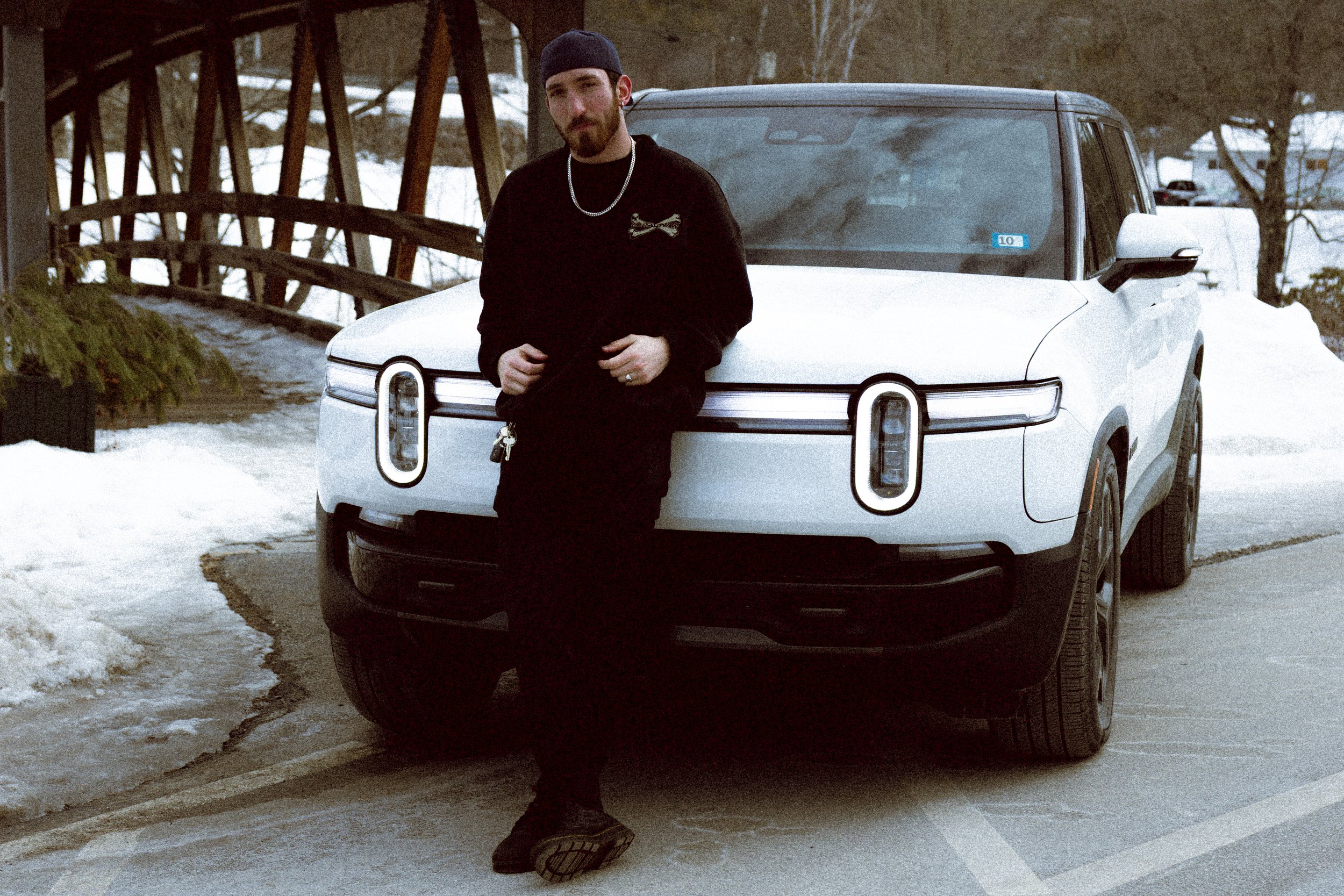 A man leaning against the front of a white electric SUV parked outdoors in a winter setting, with snow on the ground and a wooden bridge in the background.