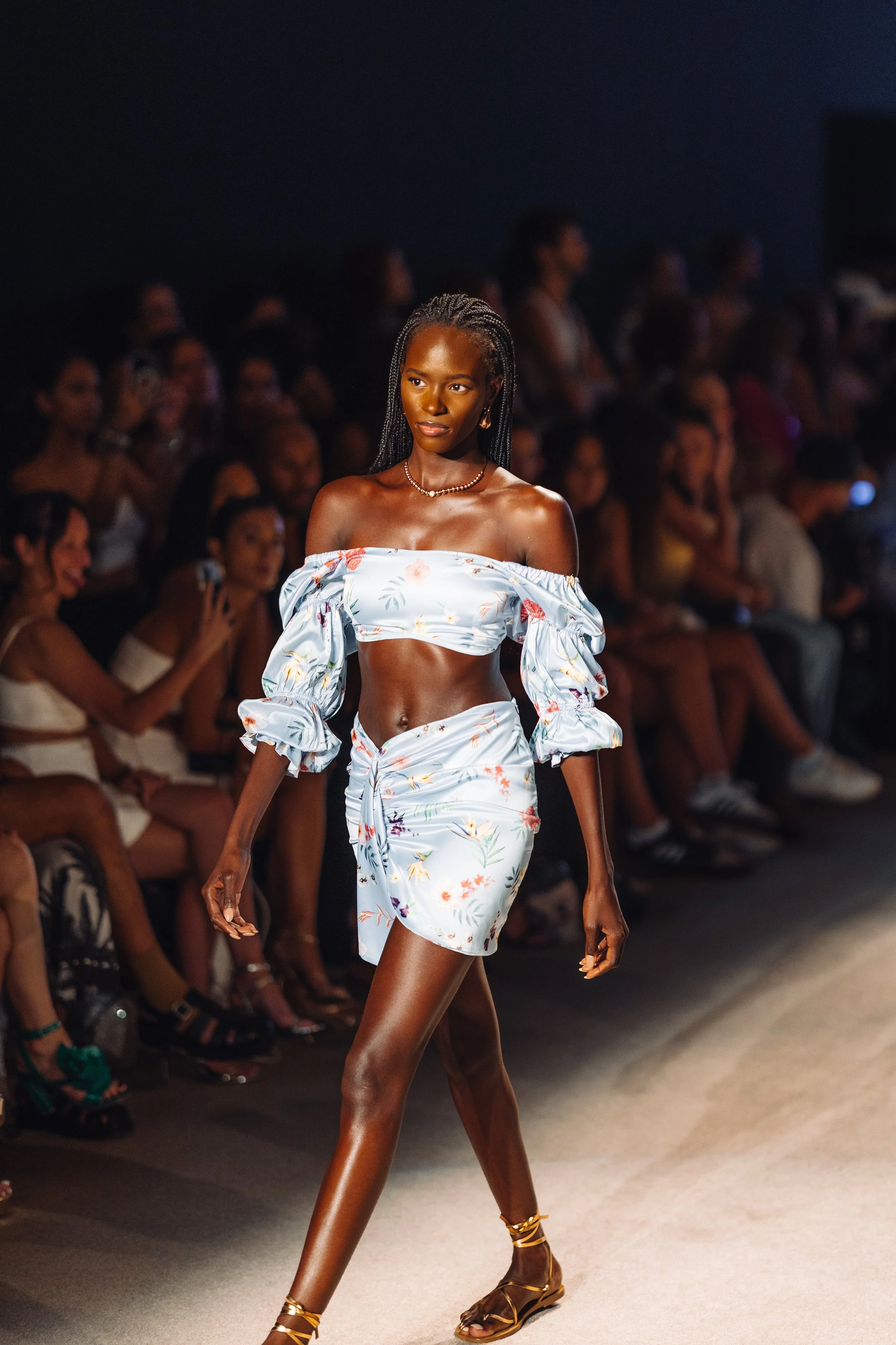 Model walking on runway in floral two-piece outfit with off-the-shoulder top and matching skirt, audience watching in background.